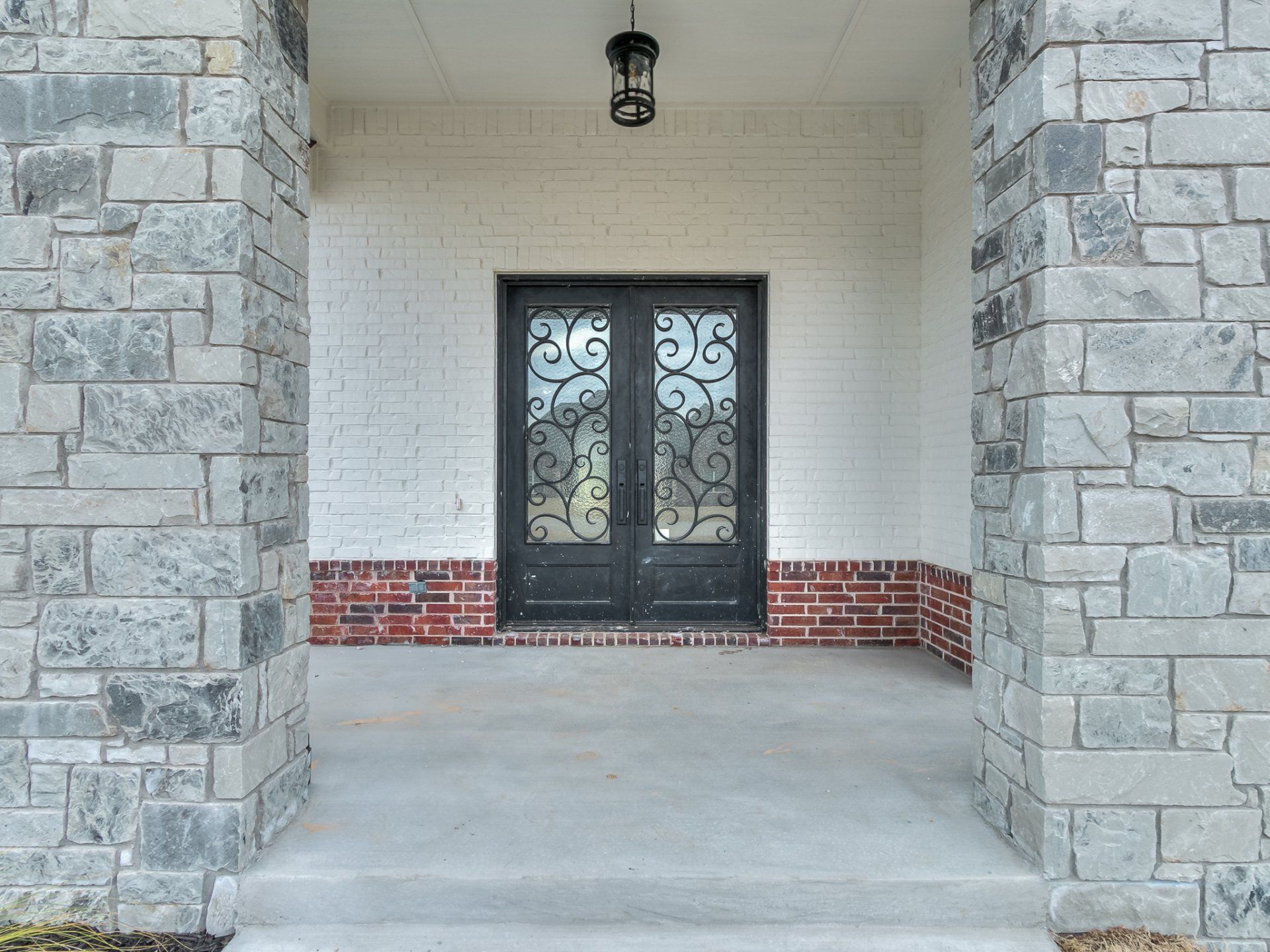 A covered porch entrance featuring stone columns, a white brick wall with a red brick base, and double ornate iron doors.