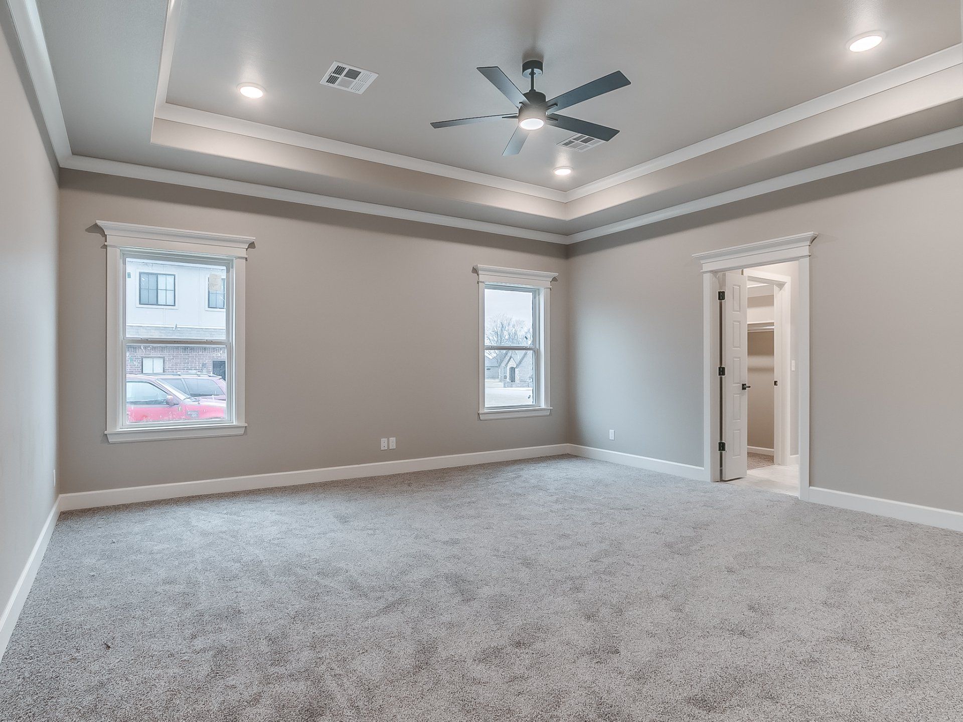Empty bedroom with grey carpet, light walls, a tray ceiling with a fan, two windows, and an open closet door.