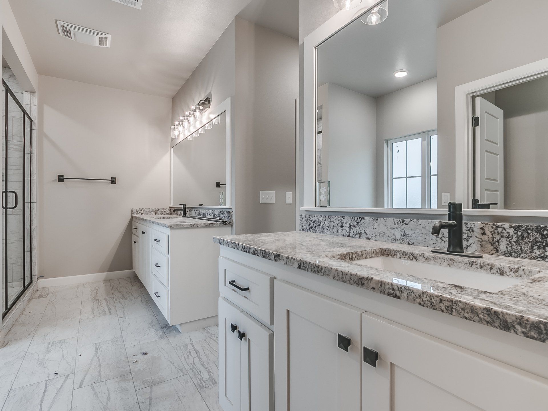A modern bathroom with white cabinets, granite countertops, dual sinks, a large mirror, and a walk-in glass shower.