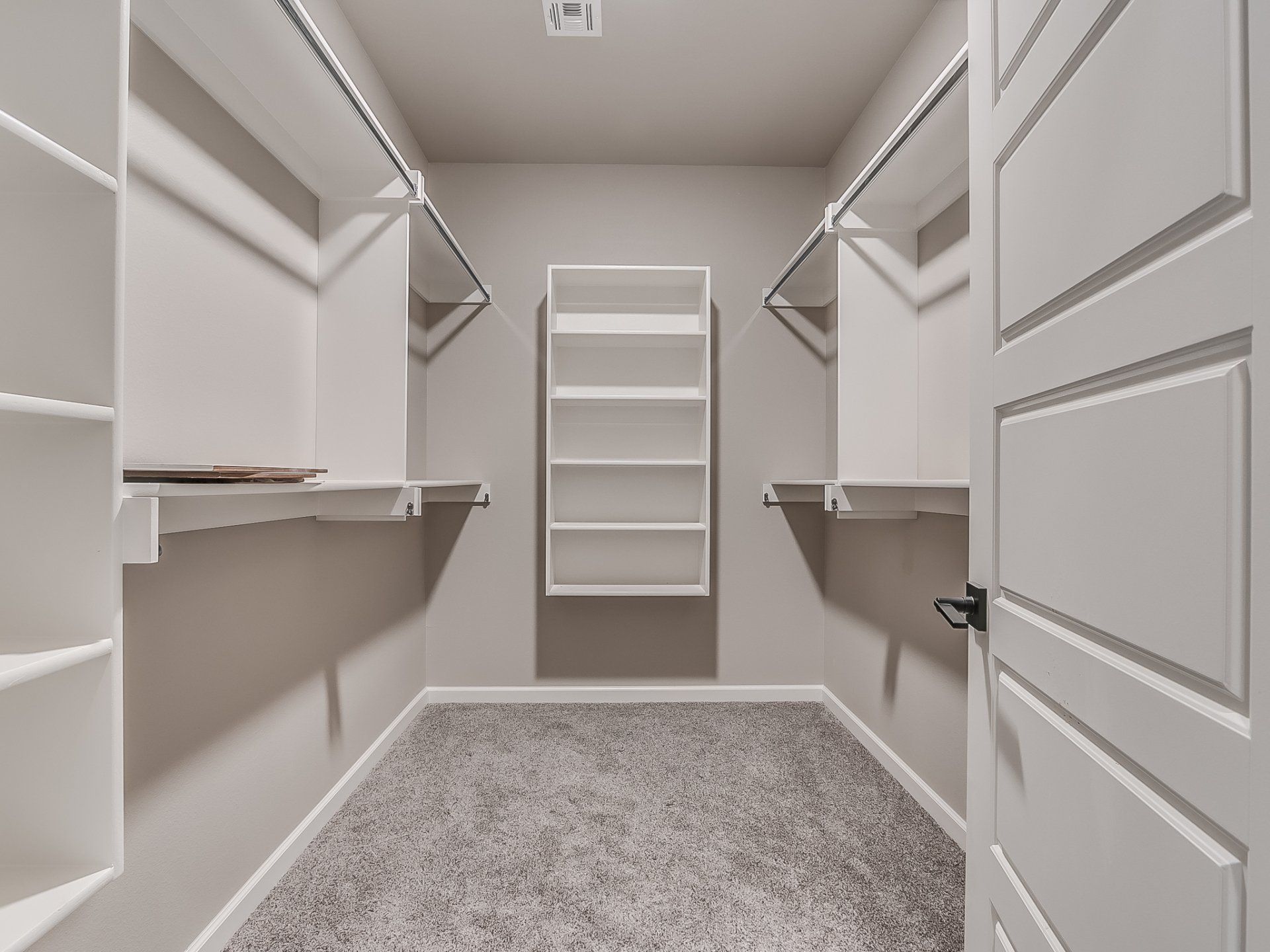 A walk-in closet with gray carpet, white shelving on both walls, a back wall shelf, and an open white door.