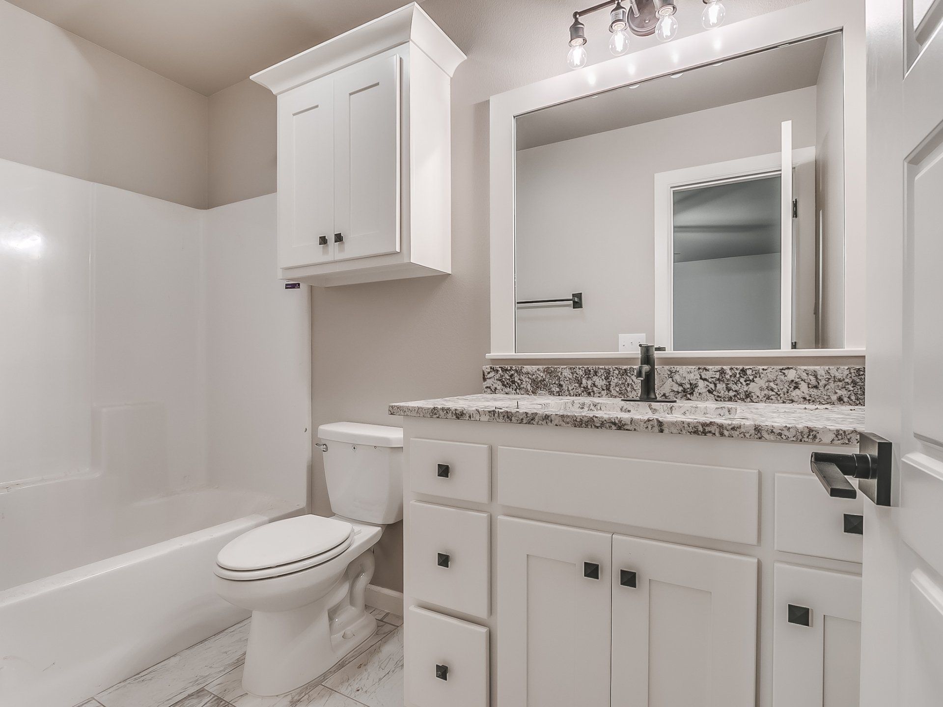 A bathroom with a white vanity, granite countertop, wall-mounted cabinet, toilet, and a white bathtub-shower combo.