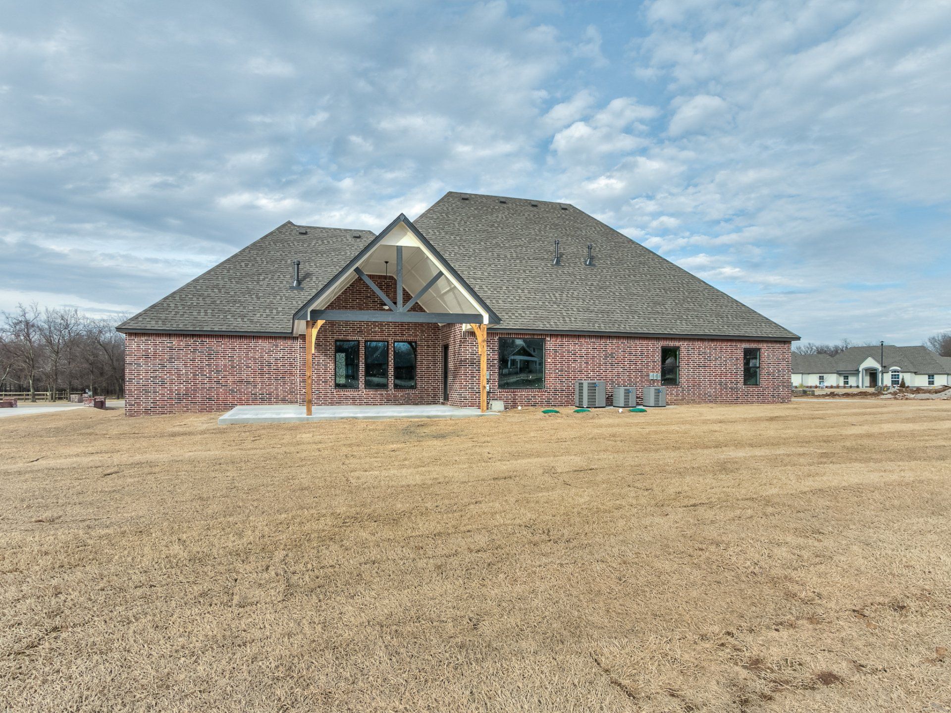 A single-story, tan brick house under construction with a large covered patio and gray shingled roof, set in a dry field.