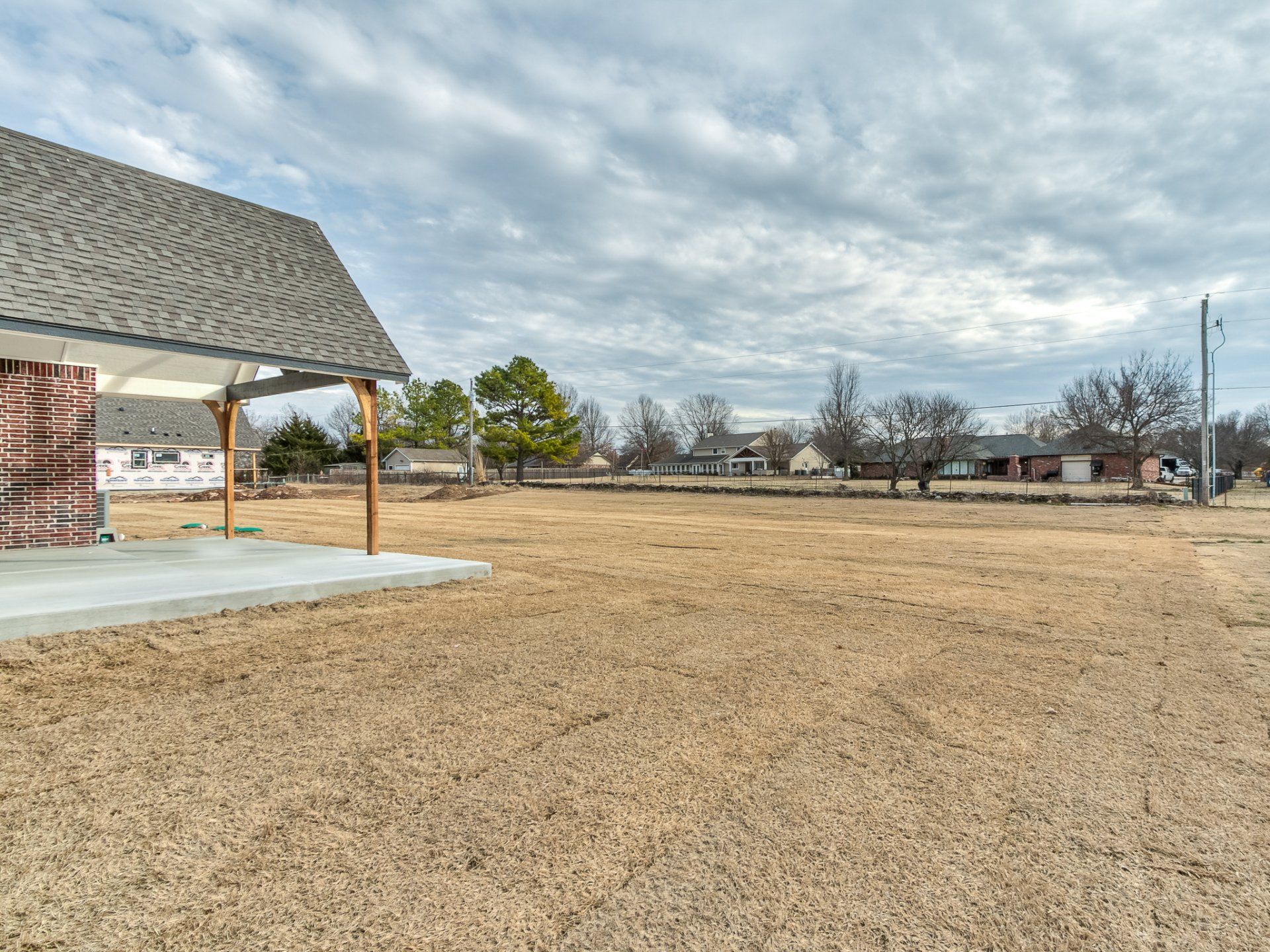 A covered patio area attached to a house overlooking a large, empty, dry grass field under a cloudy sky.