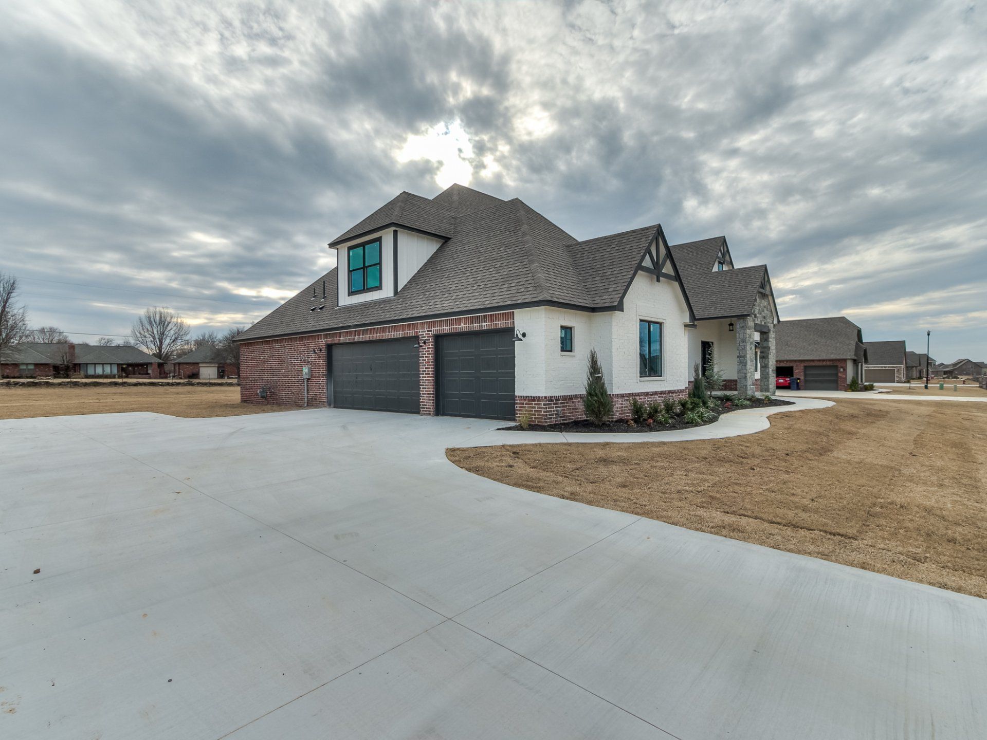A modern, multi-story house with cream stucco and stone accents, a dark shingled roof, and a large concrete driveway.