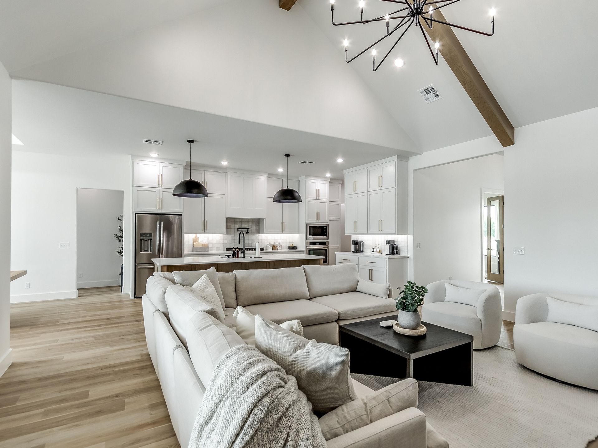 A bright, modern open-concept living room with a white sectional sofa, a black coffee table, and a kitchen in the back.