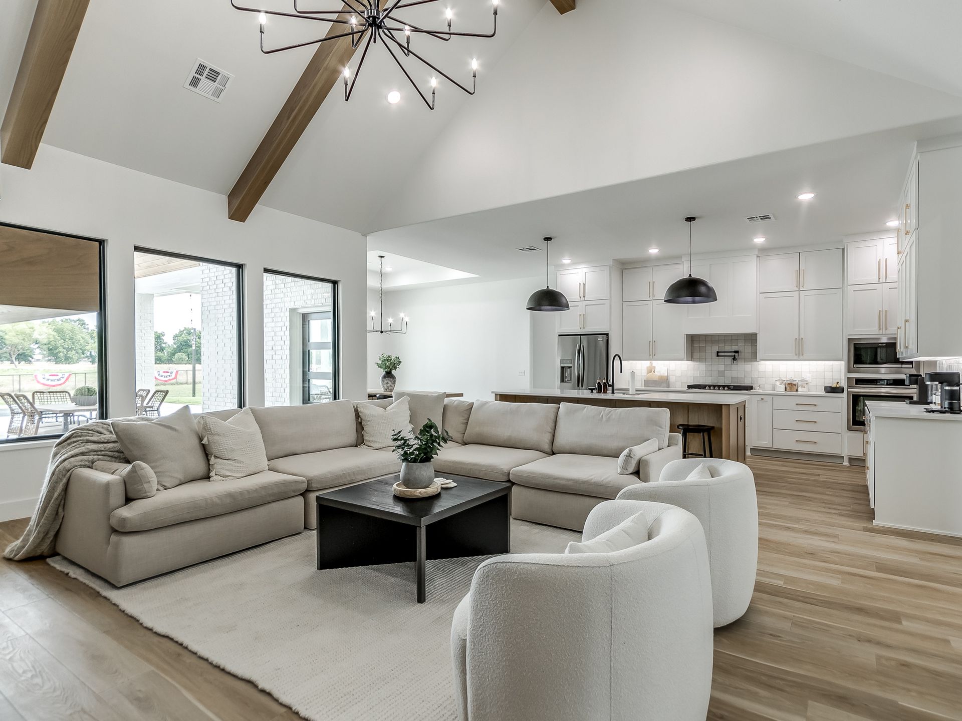 A bright, open-plan living room with a large beige sectional, two white chairs, a coffee table, and a kitchen behind it.