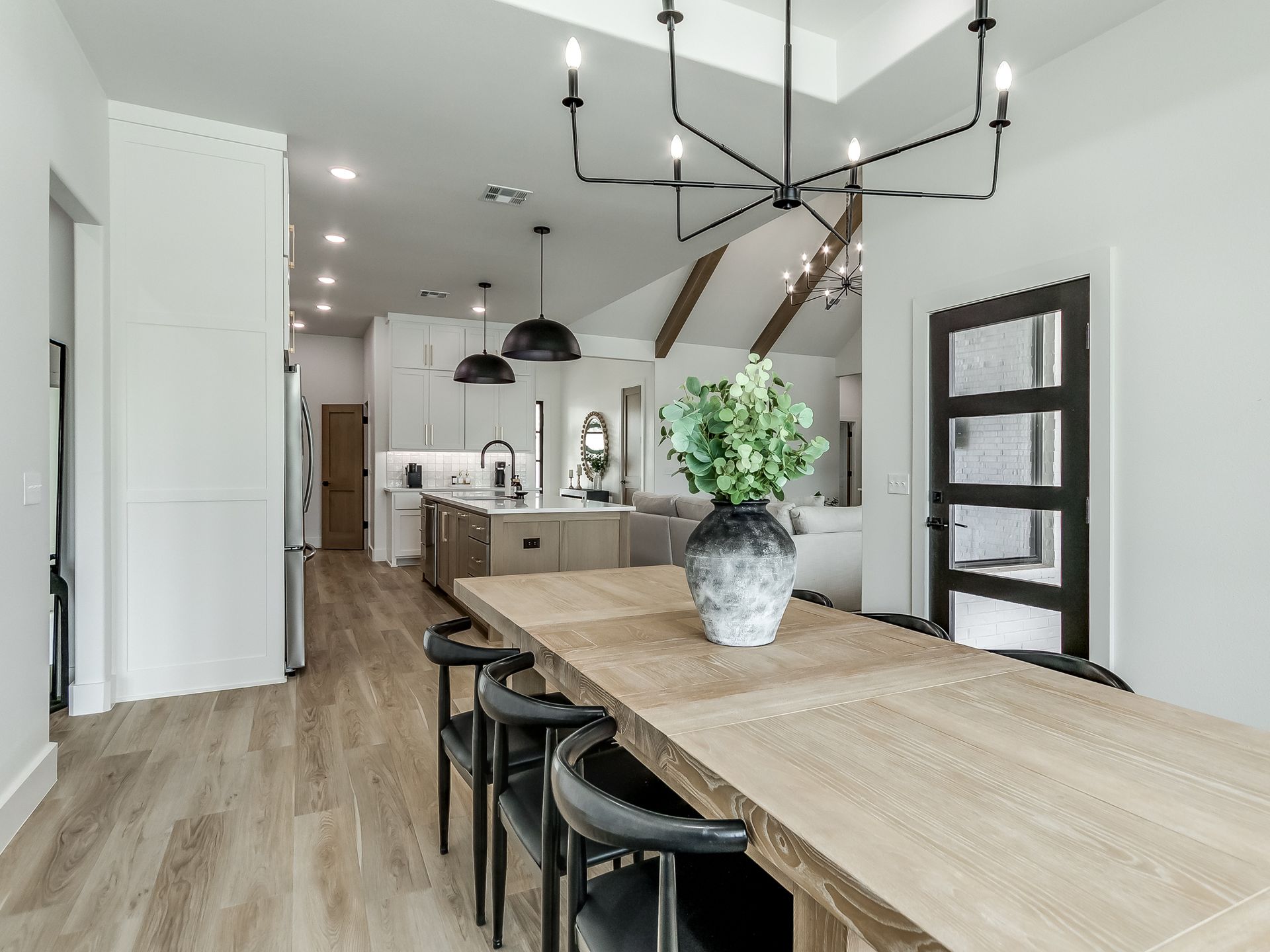 A modern dining area with a long wooden table, black chairs, and a large chandelier, leading into an open-concept kitchen.