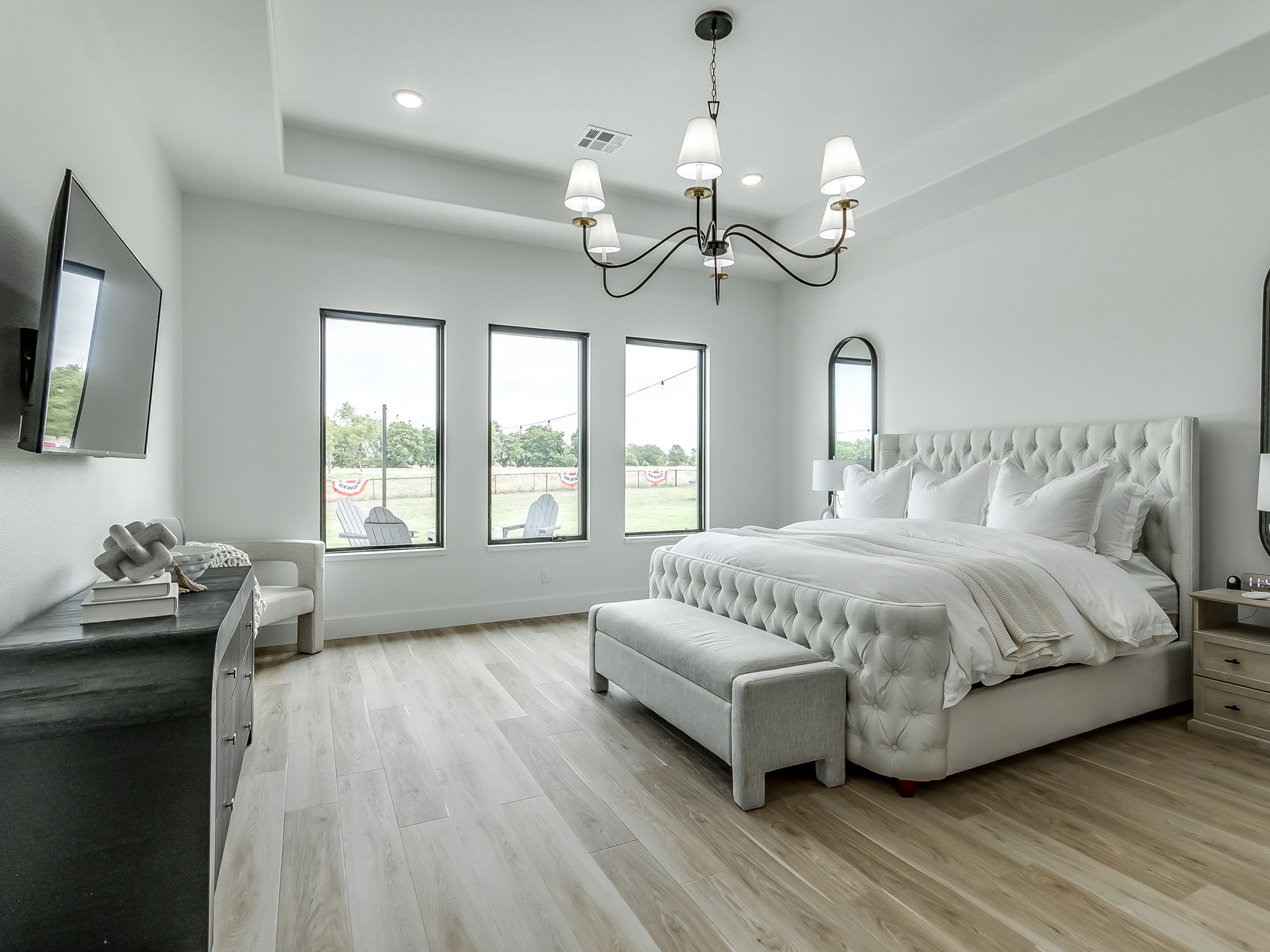 A bright, modern bedroom featuring a tufted king bed, light wood floors, a chandelier, and three large windows.