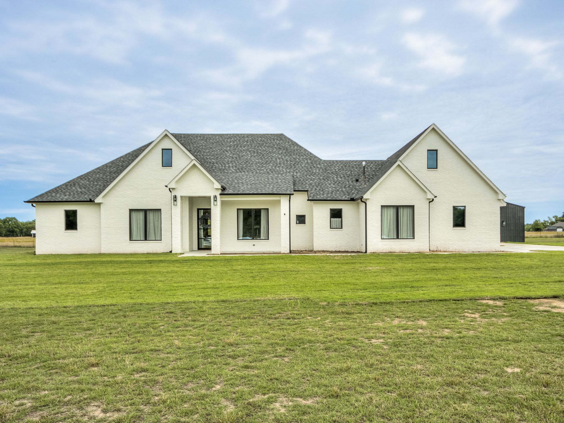 A single-story white house with a dark gray roof sits in the middle of a large green lawn under a cloudy sky.