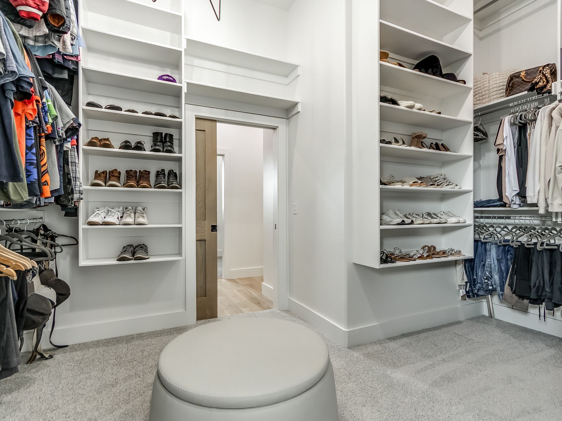 A bright, modern walk-in closet with white shelving, hanging clothes, a central light gray ottoman, and a doorway.