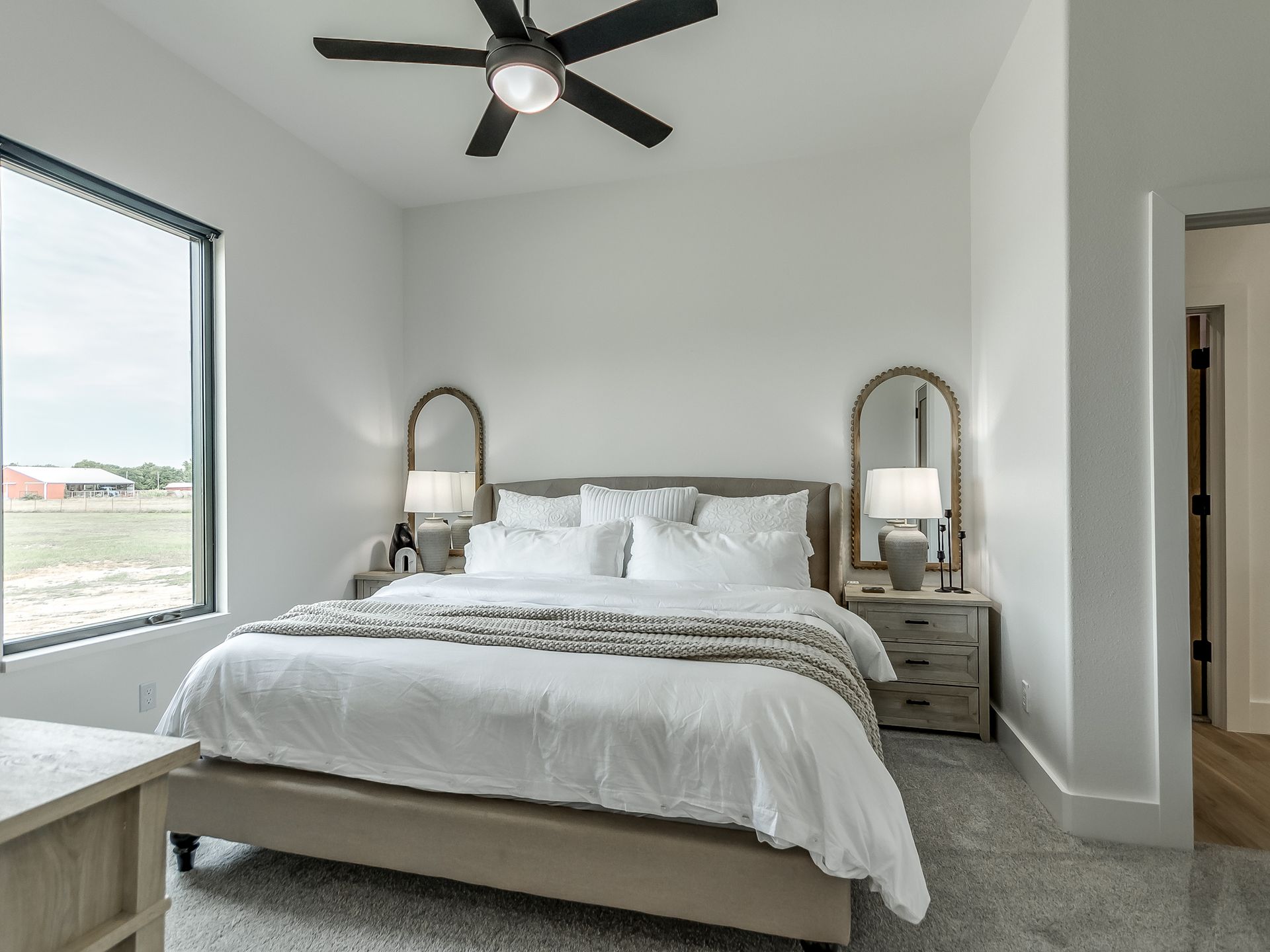 A neutral-toned bedroom with a large bed, matching nightstands with arched mirrors, and a ceiling fan by a large window.