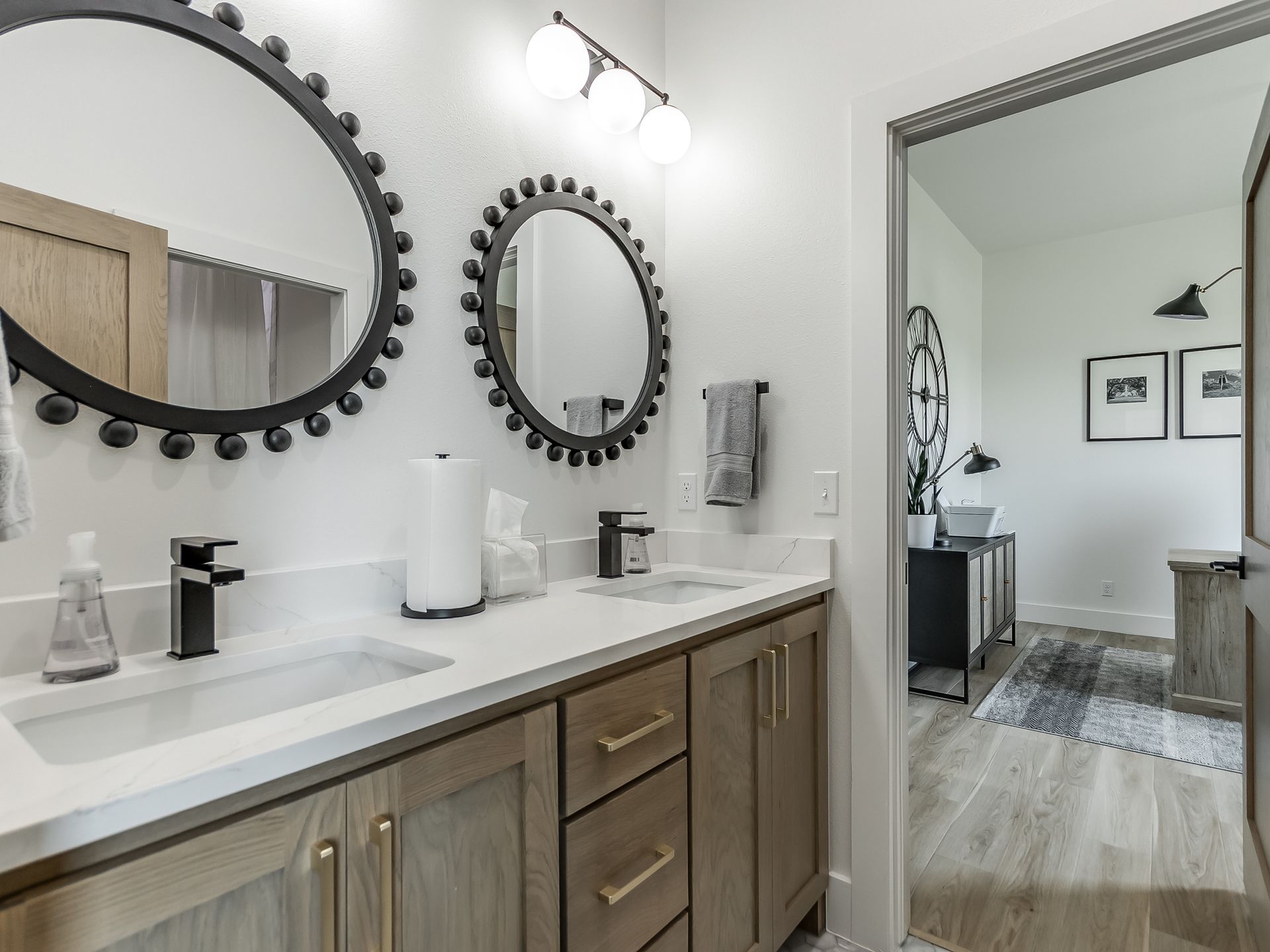 A double vanity bathroom with two round mirrors, modern lighting, wood cabinets, and an open doorway to a bedroom.