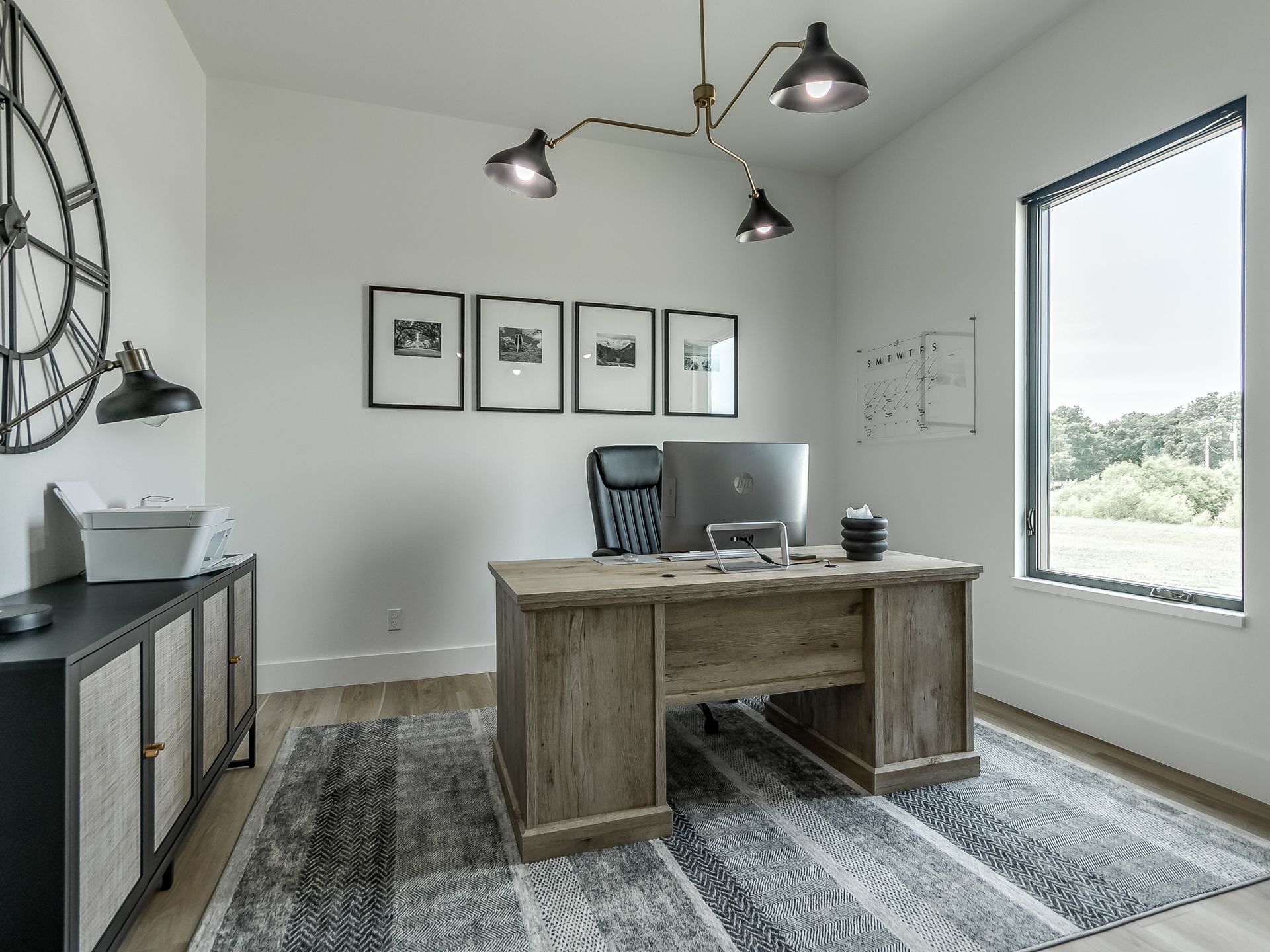 Modern home office with a wooden desk, rolling chair, printer, large wall clock, and a window overlooking greenery.