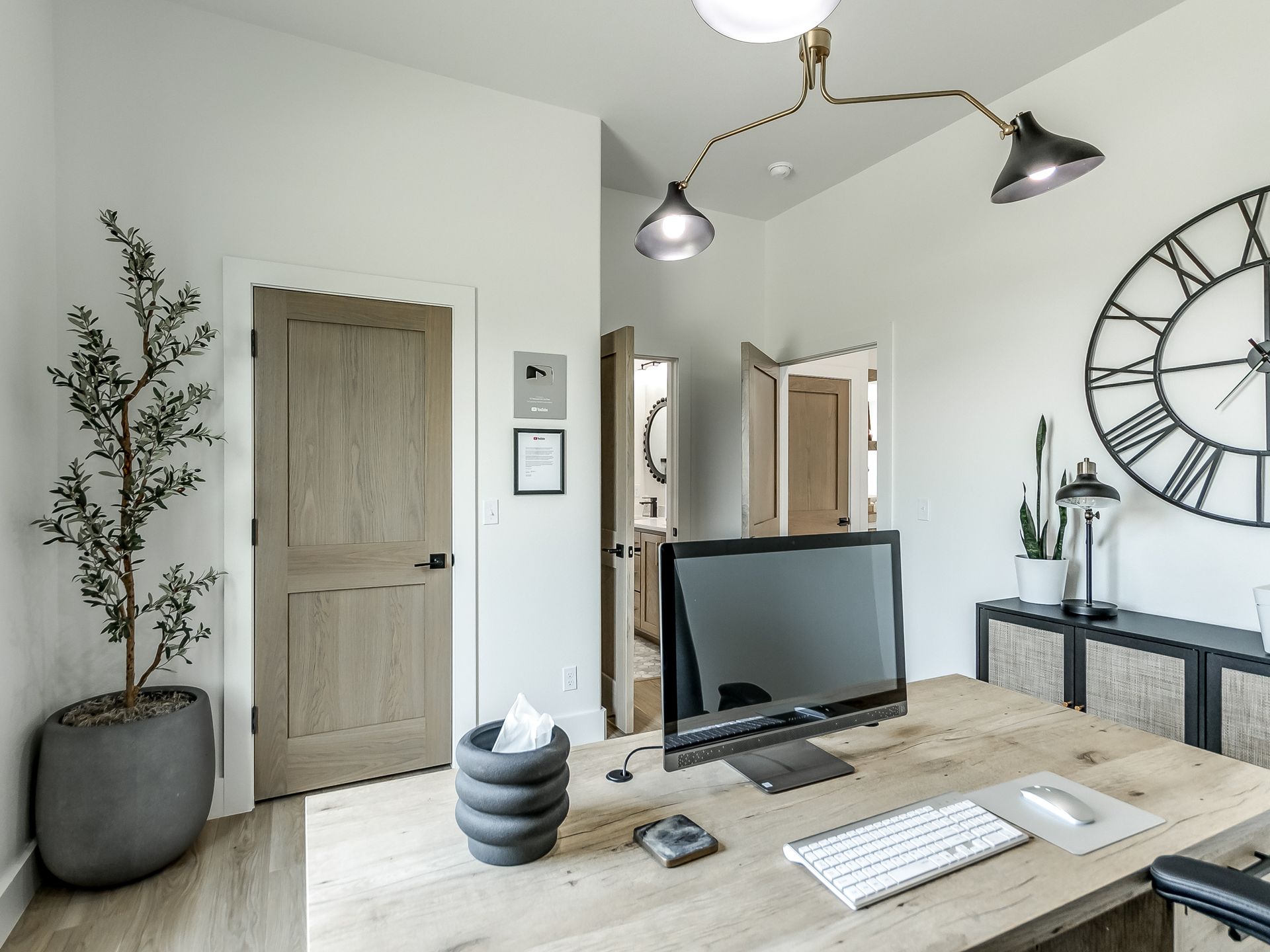 A modern, minimalist home office with a light wood desk, computer, decorative wall clock, and potted plant.