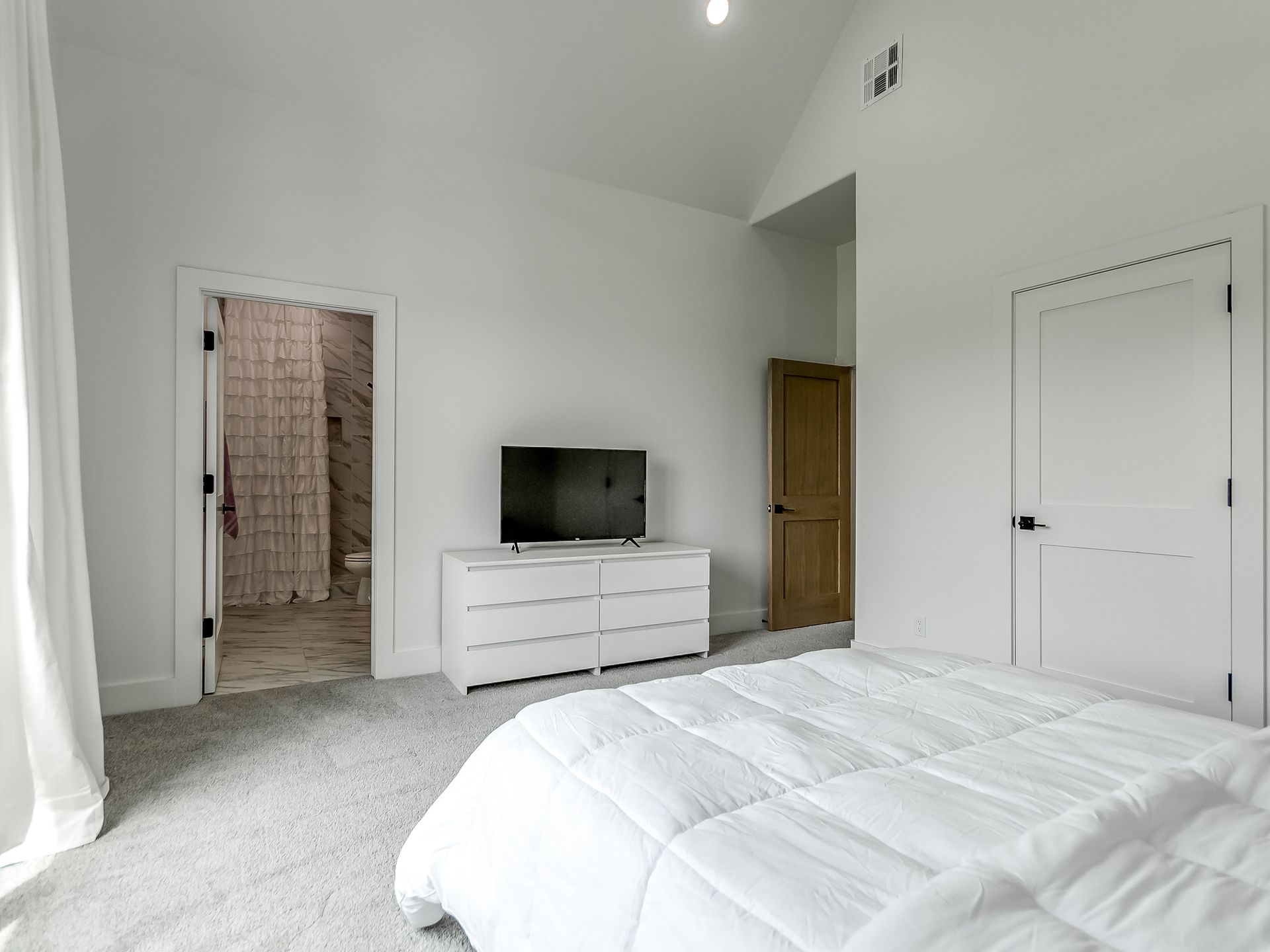 A bright bedroom with white walls, carpeted floors, a white dresser with a TV, and doors to a bathroom and closet.