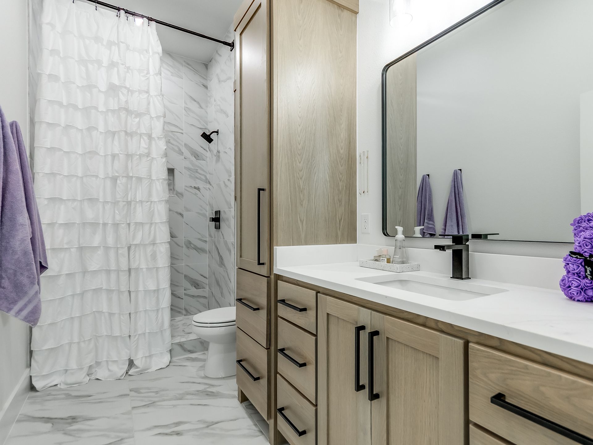 A modern bathroom featuring a wood vanity, marble-style flooring and shower walls, and a ruffled white shower curtain.