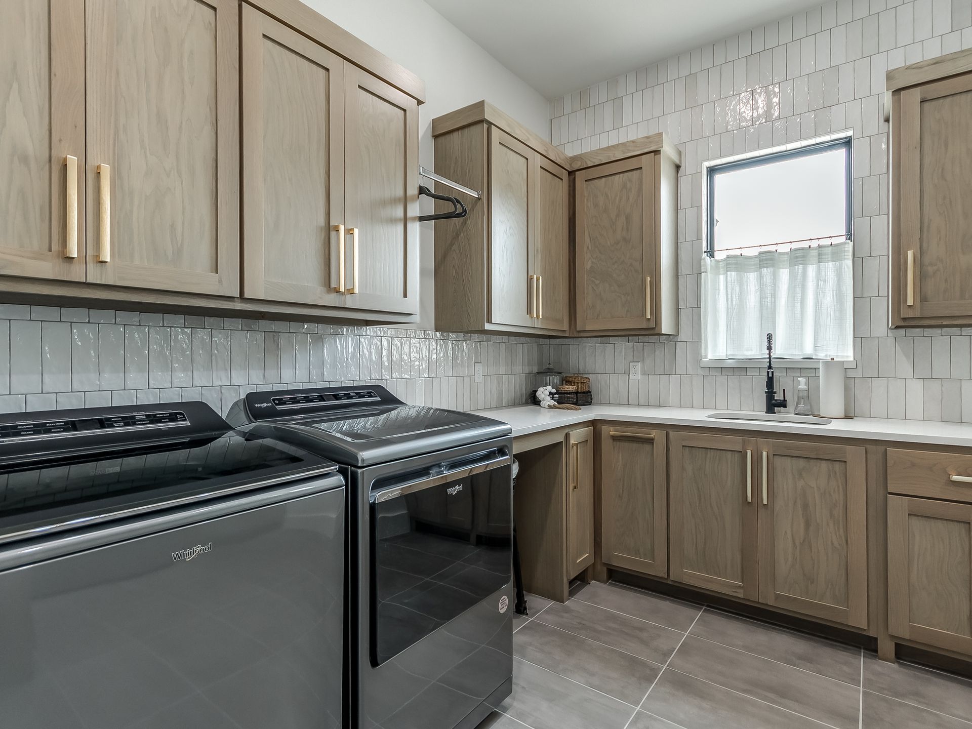A modern laundry room with light wood cabinets, gray appliances, a tiled backsplash, and a window above a utility sink.