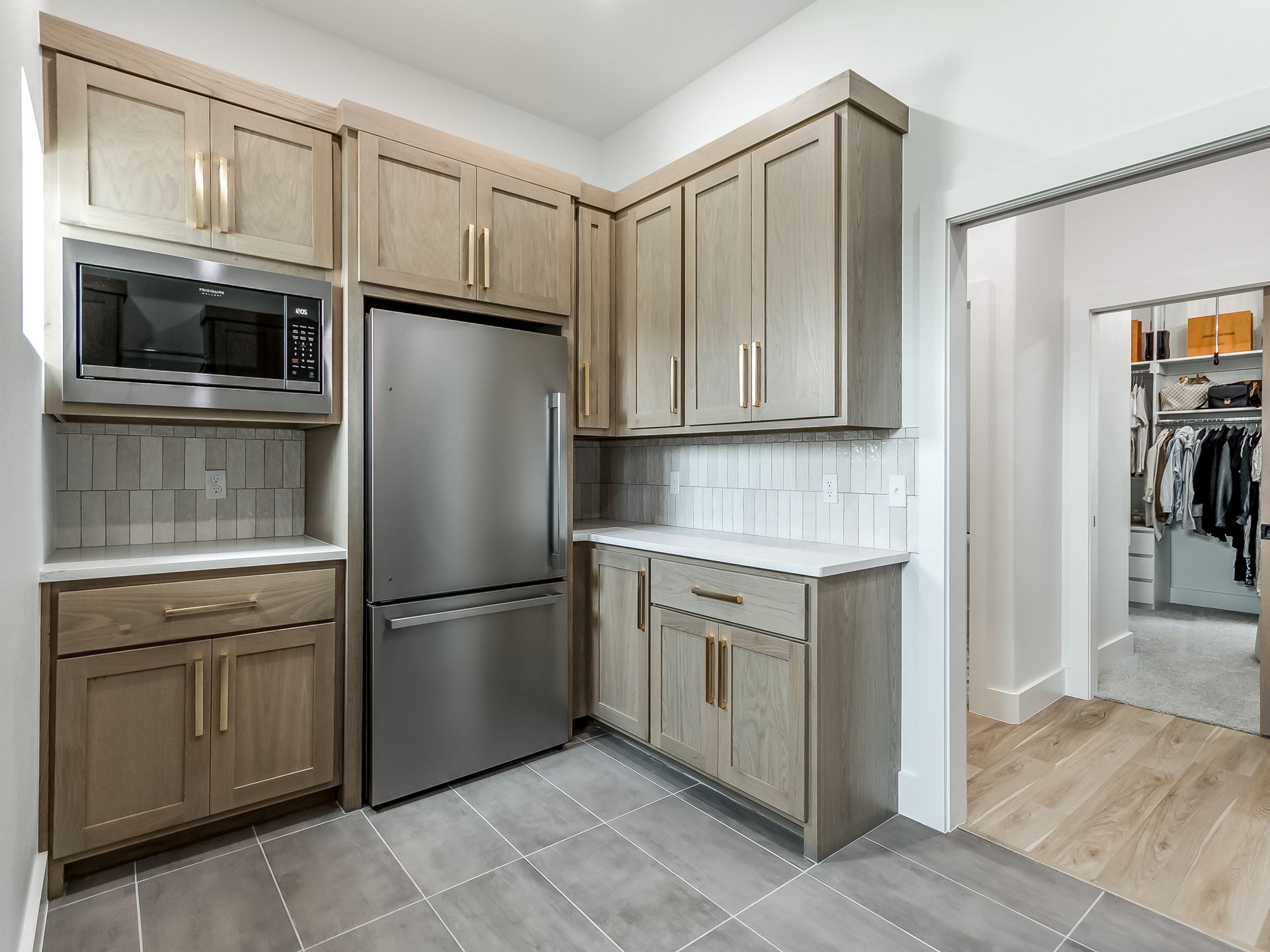 A modern kitchen area with light wood cabinets, stainless steel appliances, and gray tiled floors leading to a walk-in closet.