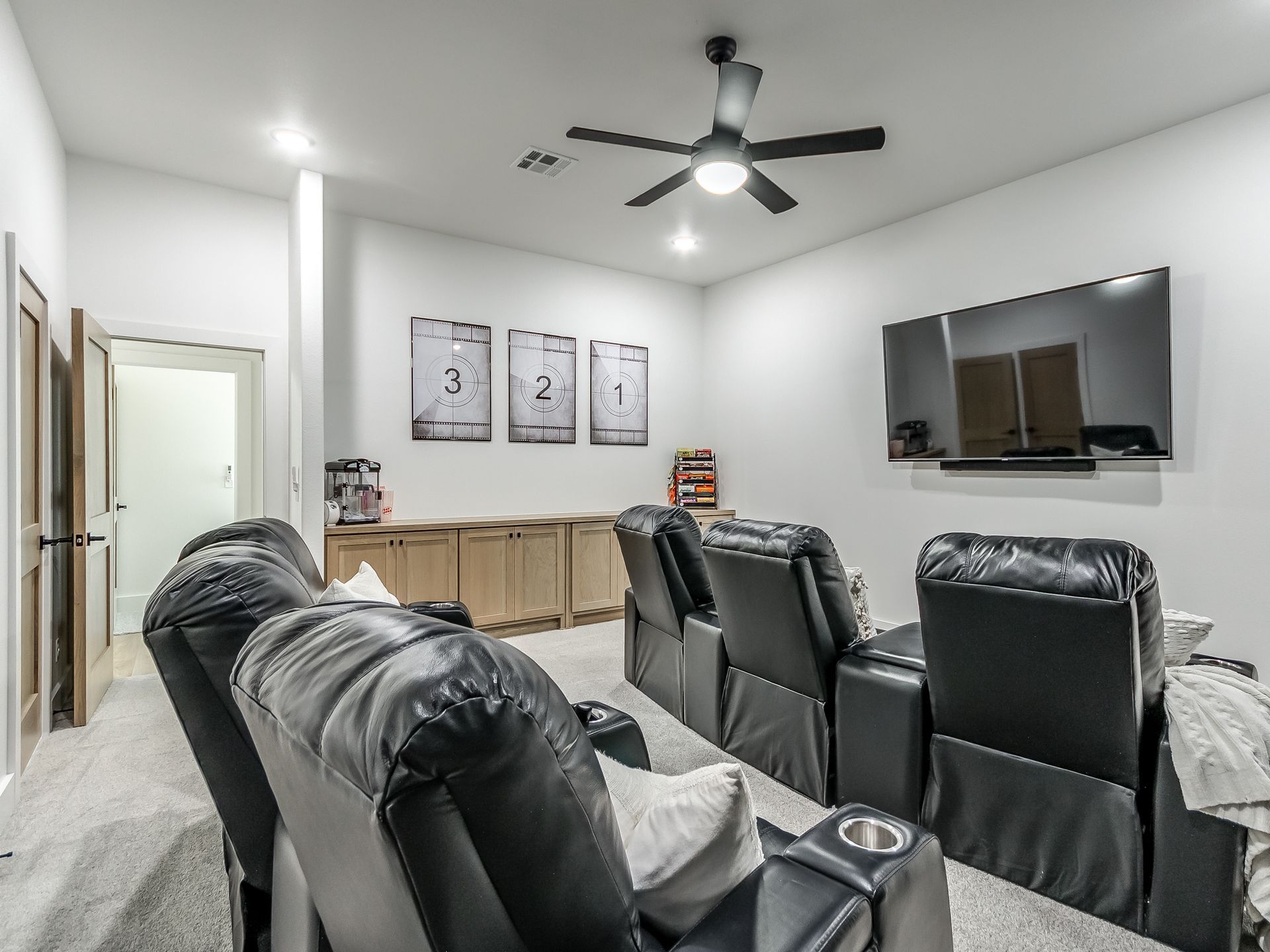 A home theater room with black leather reclining chairs, wall-mounted television, and framed numbered art prints.