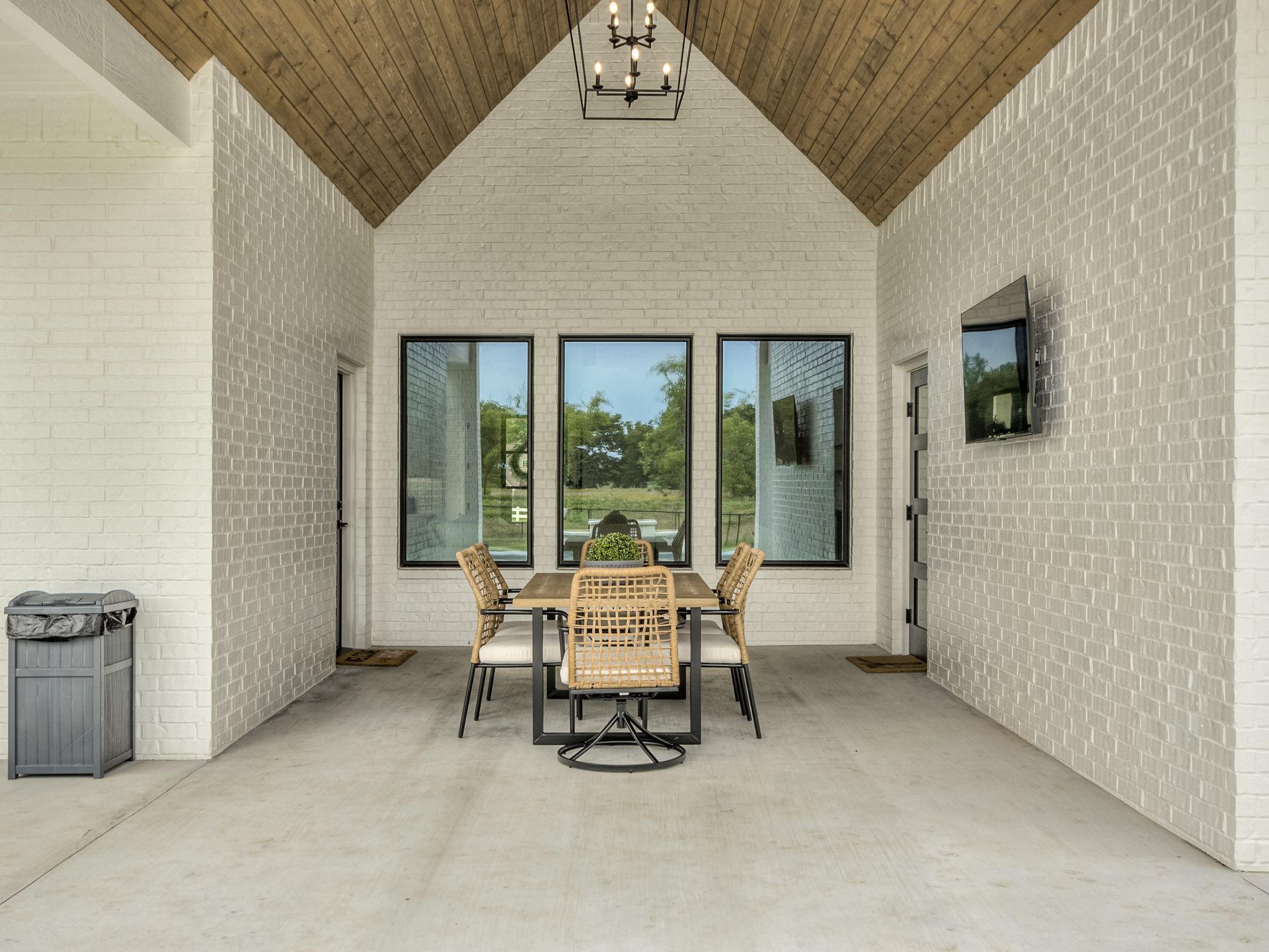 A covered outdoor dining area with a rustic wooden ceiling, white brick walls, a wooden table, and woven chairs.