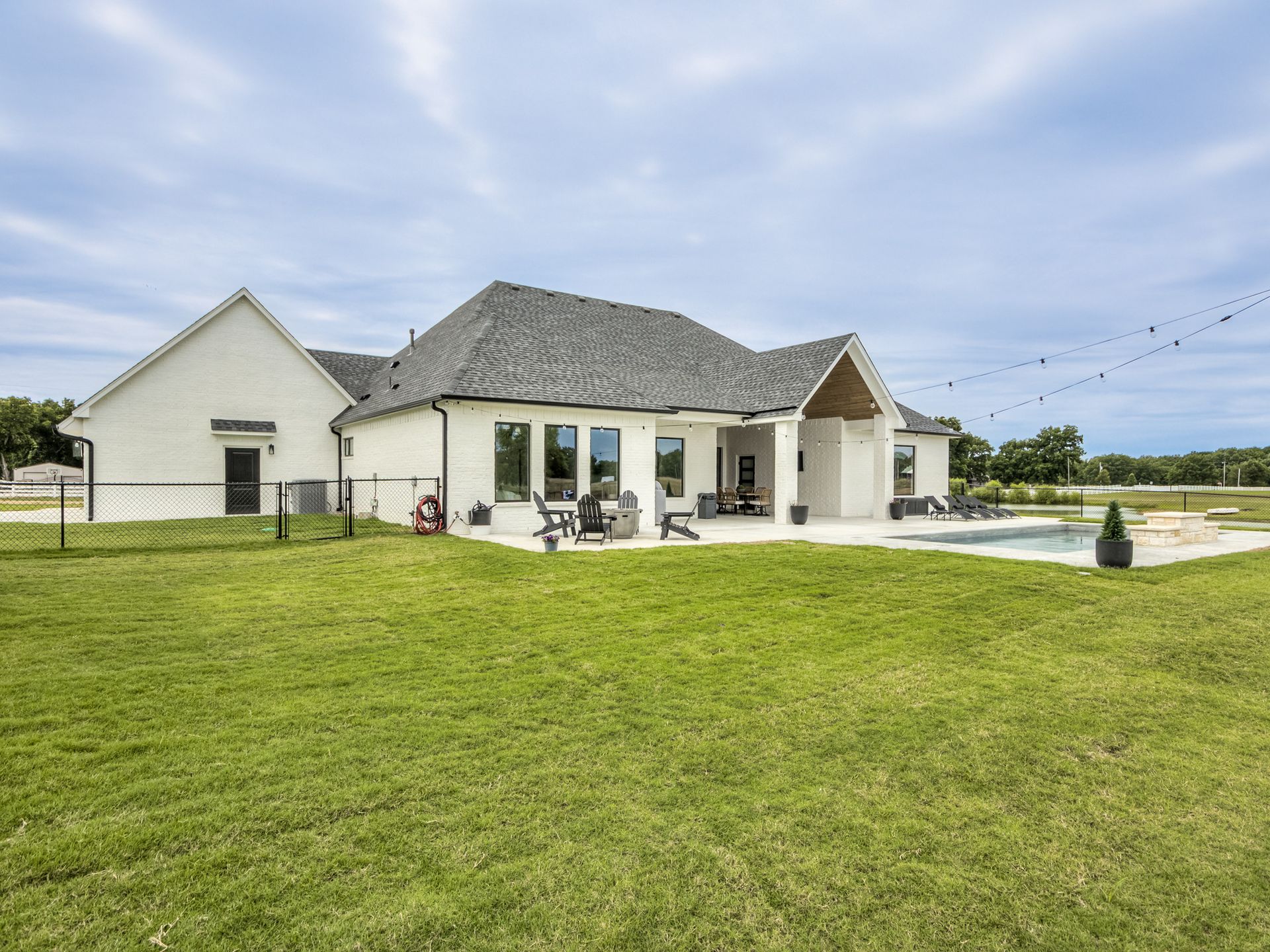 Back exterior of a white modern house with a gray roof, lawn, patio, and a swimming pool under a partly cloudy sky.