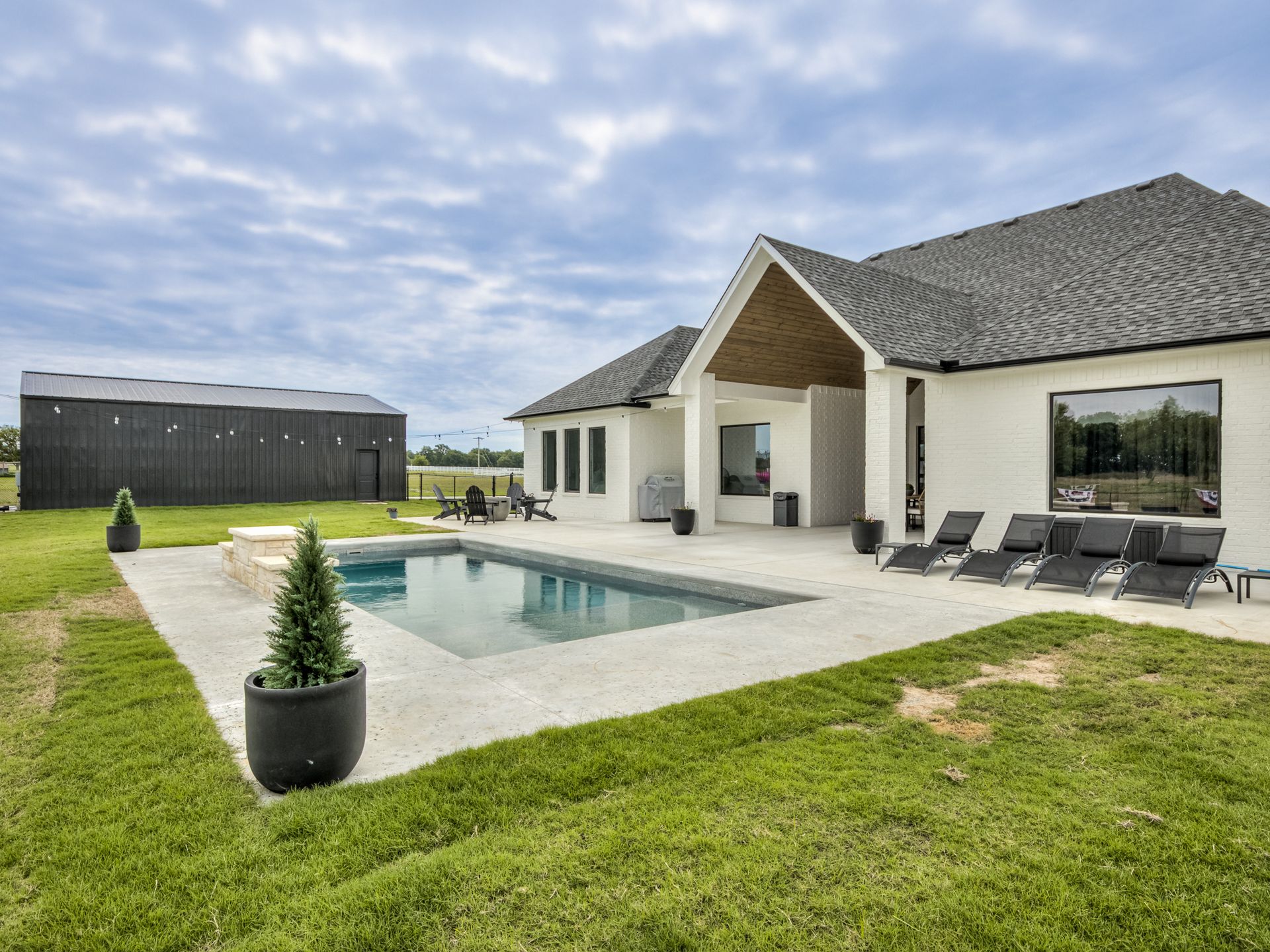 A white house with a dark roof, pool, and patio, alongside a black outbuilding under a cloudy sky.