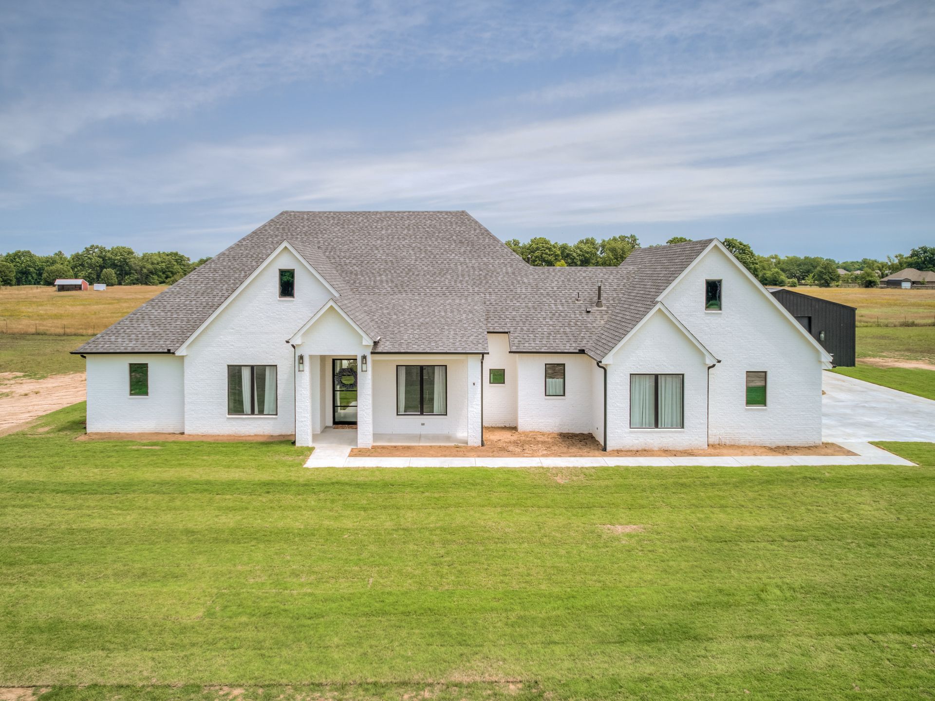 A single-story white house with a grey roof and front porch, set in a rural landscape with green grass and a blue sky.