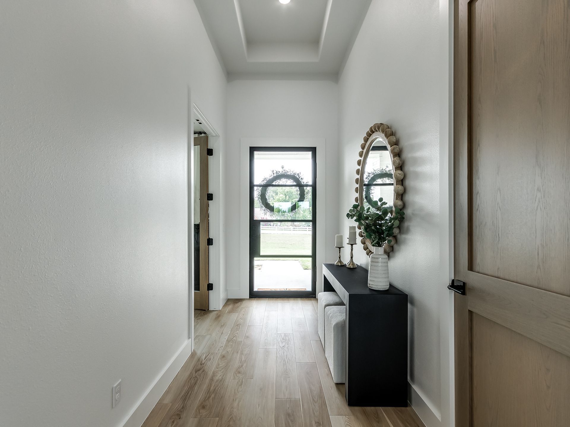A bright entryway with wood floors, a black console table, a round mirror, and a glass front door leading to a garden.