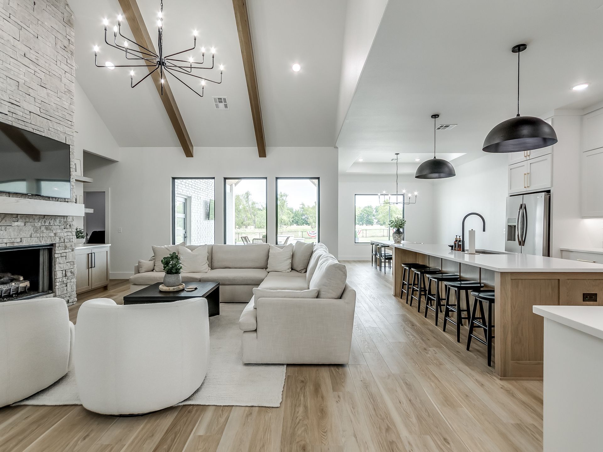 Bright, modern open-concept living room and kitchen with white furniture, stone fireplace, and wood-beam vaulted ceiling.