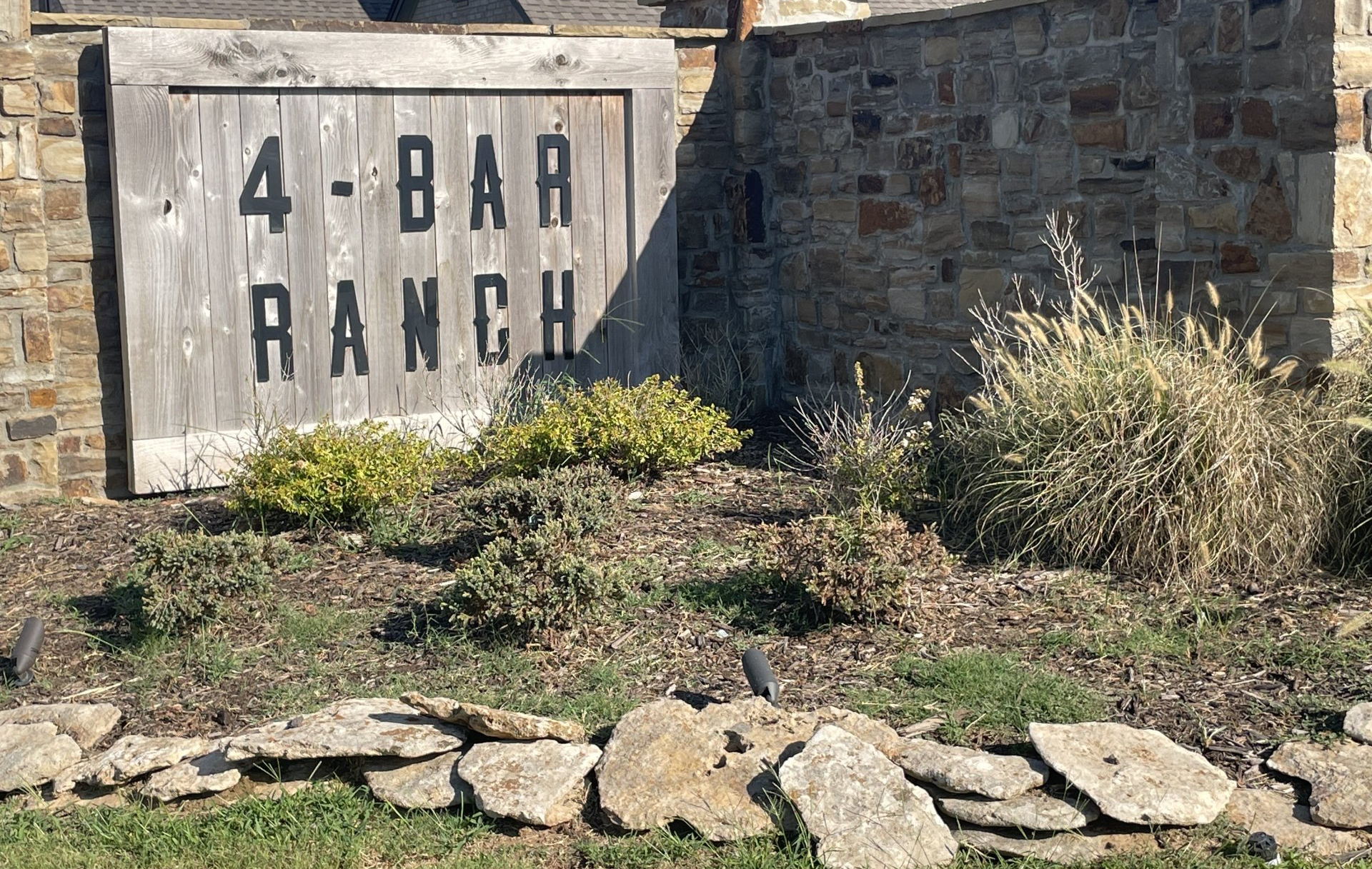 A stone-walled monument sign for 4-Bar Ranch, featuring a weathered wooden board with the name in bold black letters.
