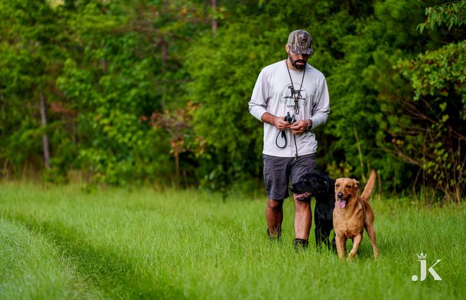 A person in a light long-sleeve shirt and cap walks through a grassy field with a black dog and a tan dog.