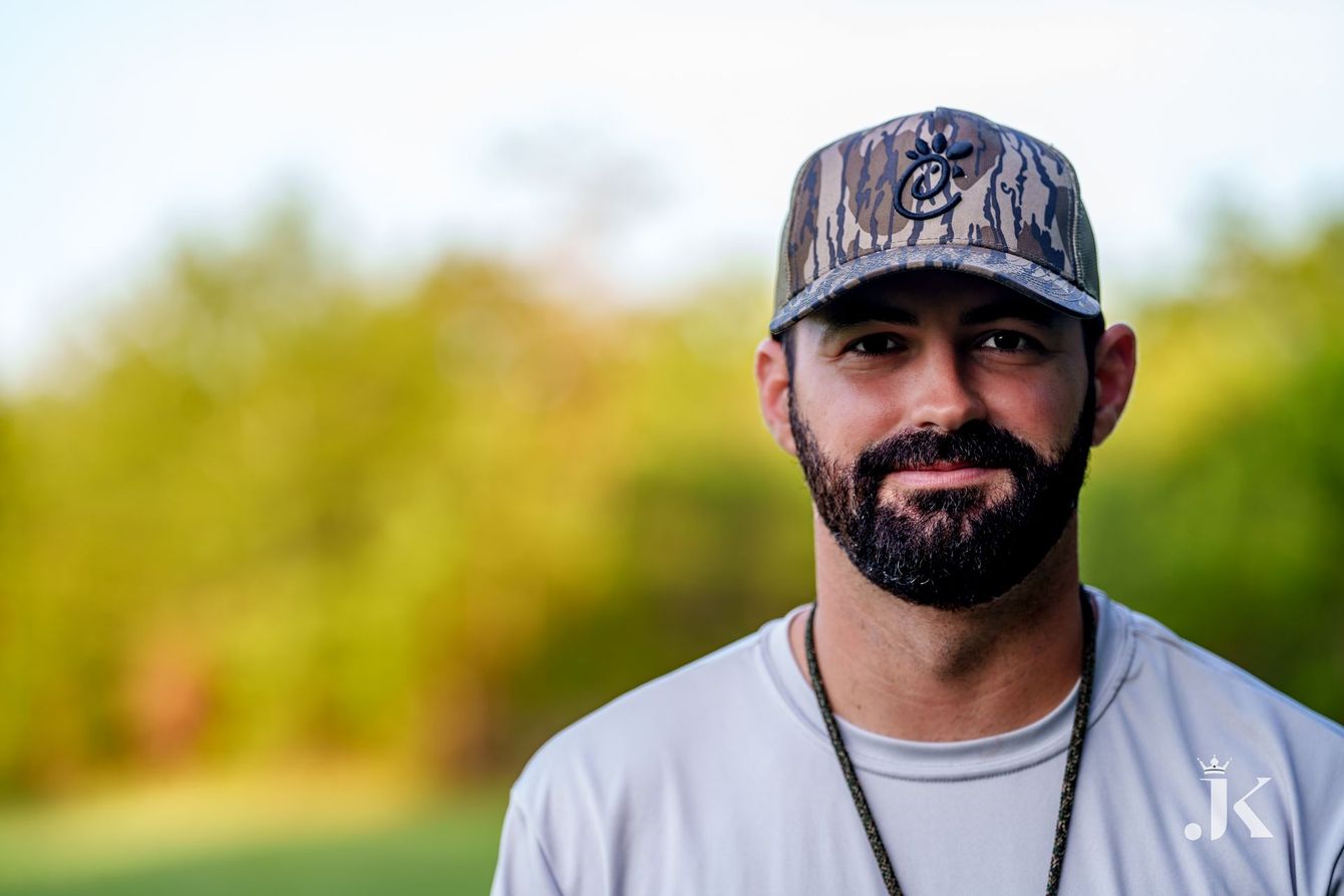 A person with a beard wearing a camouflage baseball cap and a gray t-shirt, posed against a soft-focus, blurred landscape.