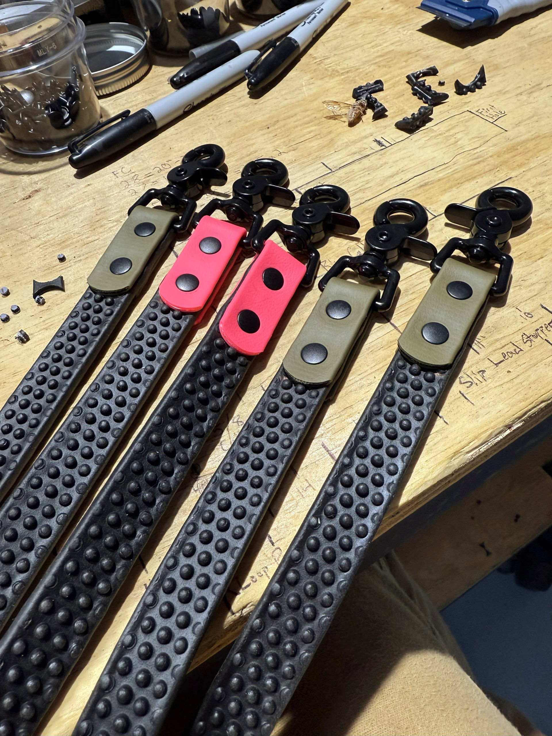 Five tactile, textured black straps with black hardware, featuring tan or bright pink accent tabs, laid on a workbench.