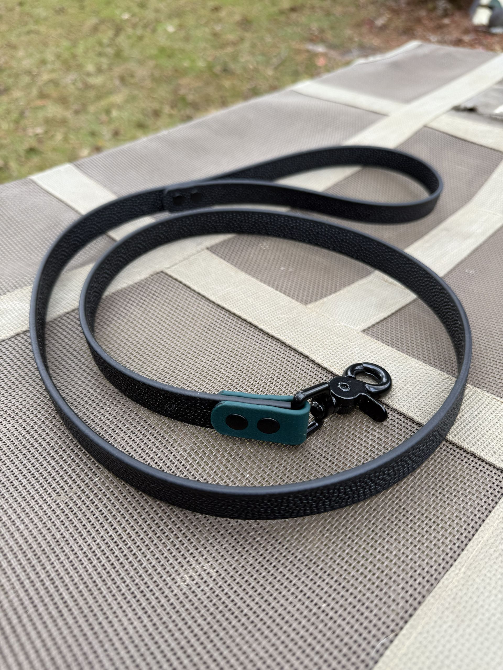 A black textured dog leash with a dark green connector and a black metal clasp, resting on a beige outdoor table.