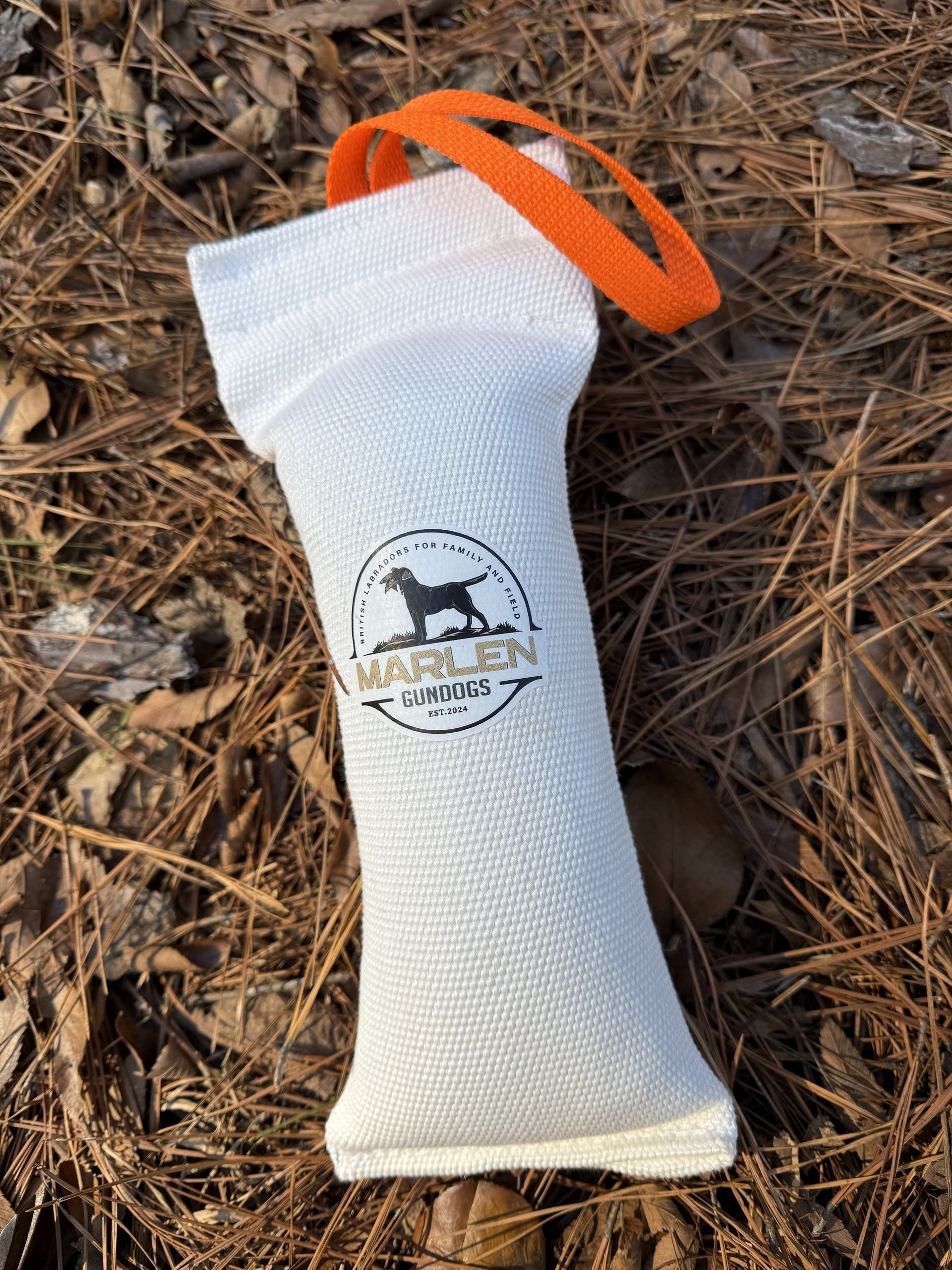 A white canvas dog training bumper with an orange handle and the MArlen logo, lying on a bed of dry pine needles.