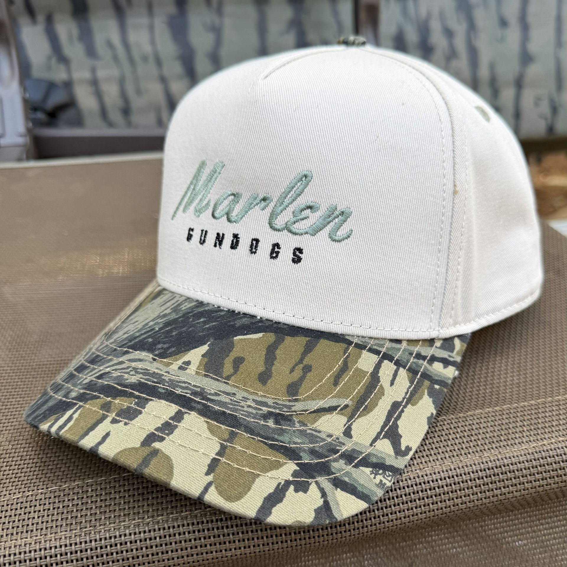 A white baseball cap with a camo-patterned brim, featuring green embroidered