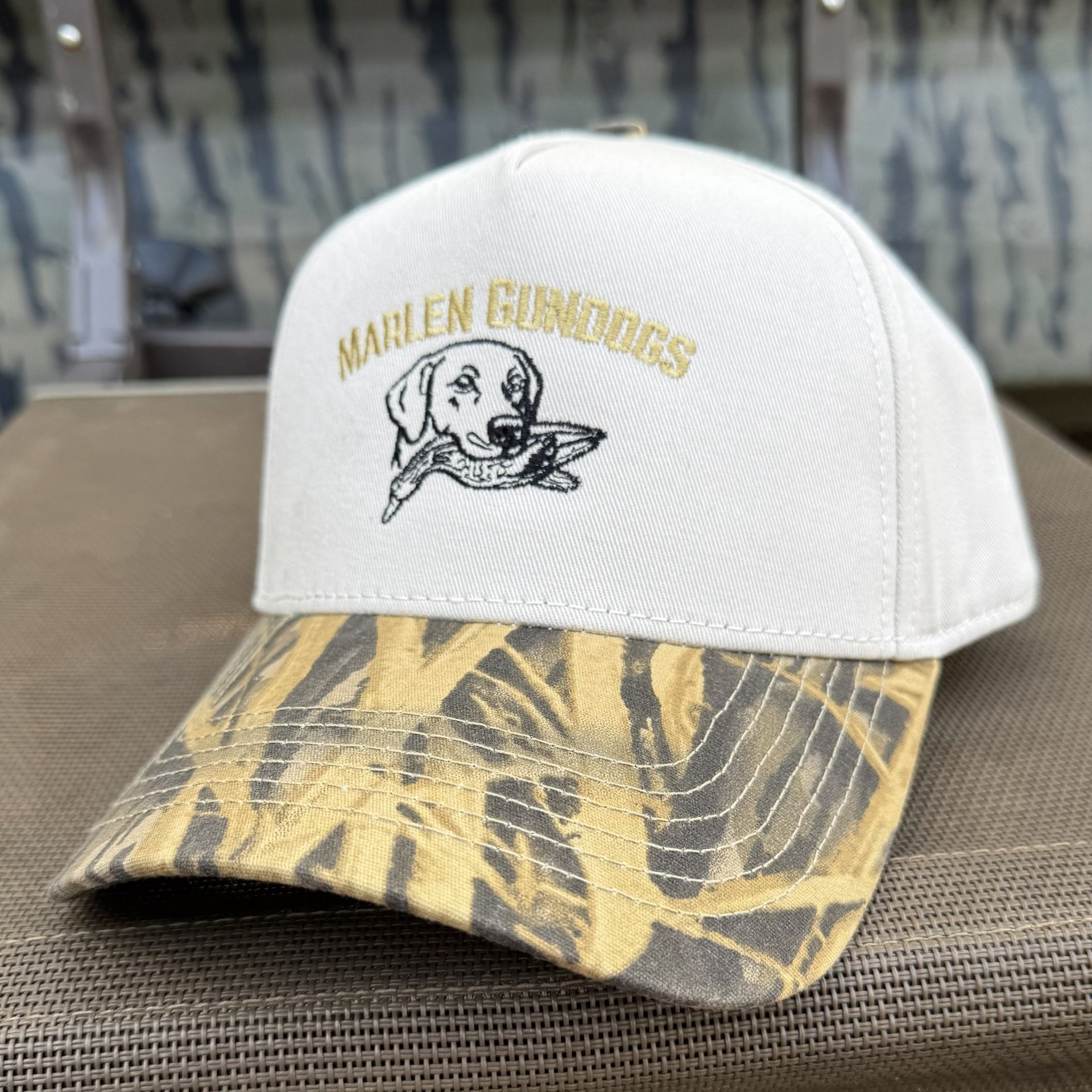 A white trucker hat with a camouflage brim, featuring an embroidered graphic of a dog holding a duck and gold text.