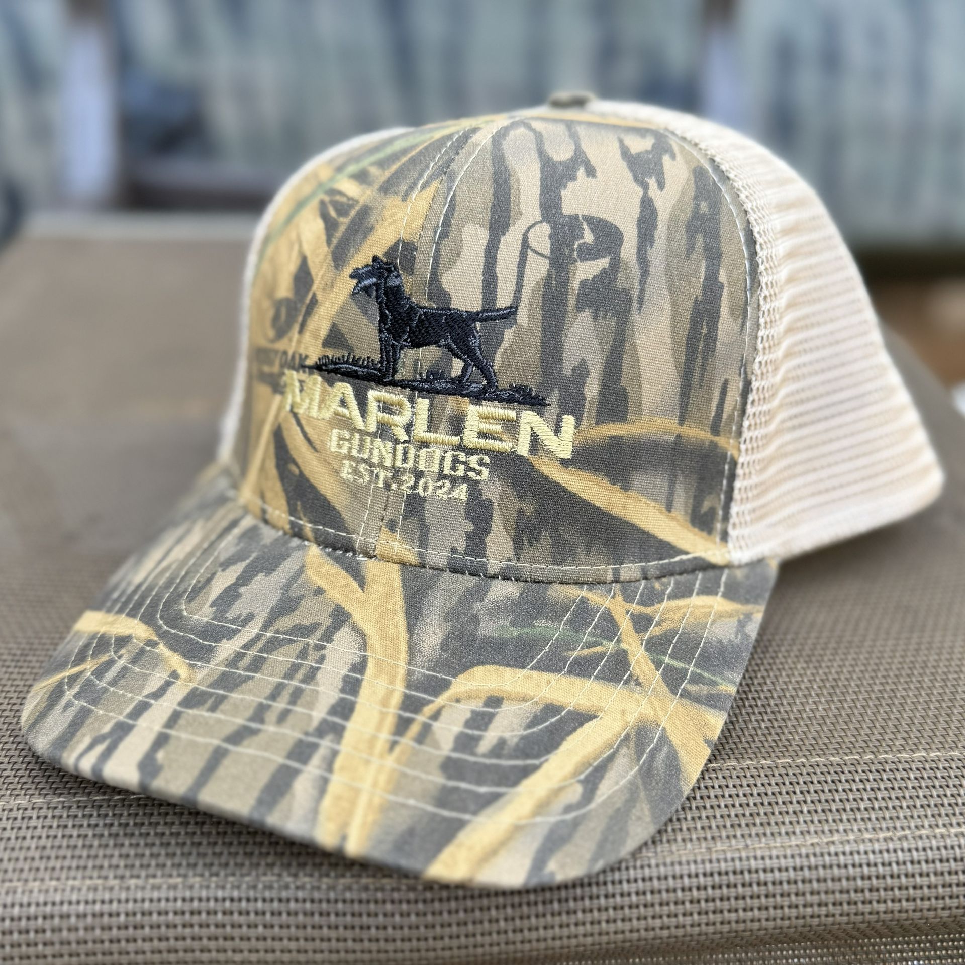 A camouflage trucker hat with a white mesh back featuring an embroidered black dog logo and the text