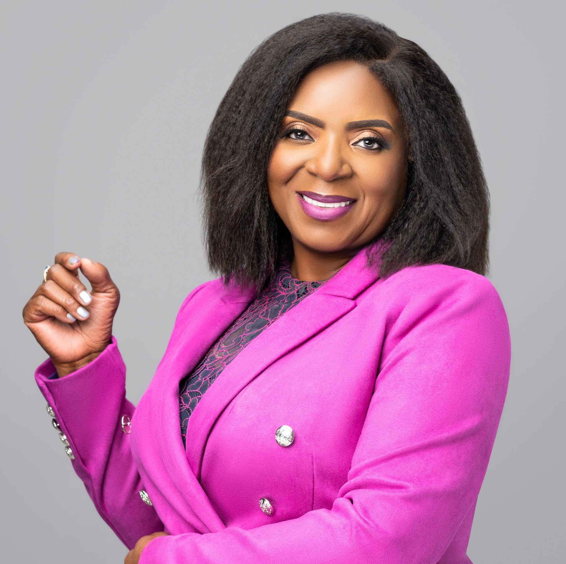 Woman in pink blazer, smiling, gesturing, with short black hair, against a gray background.