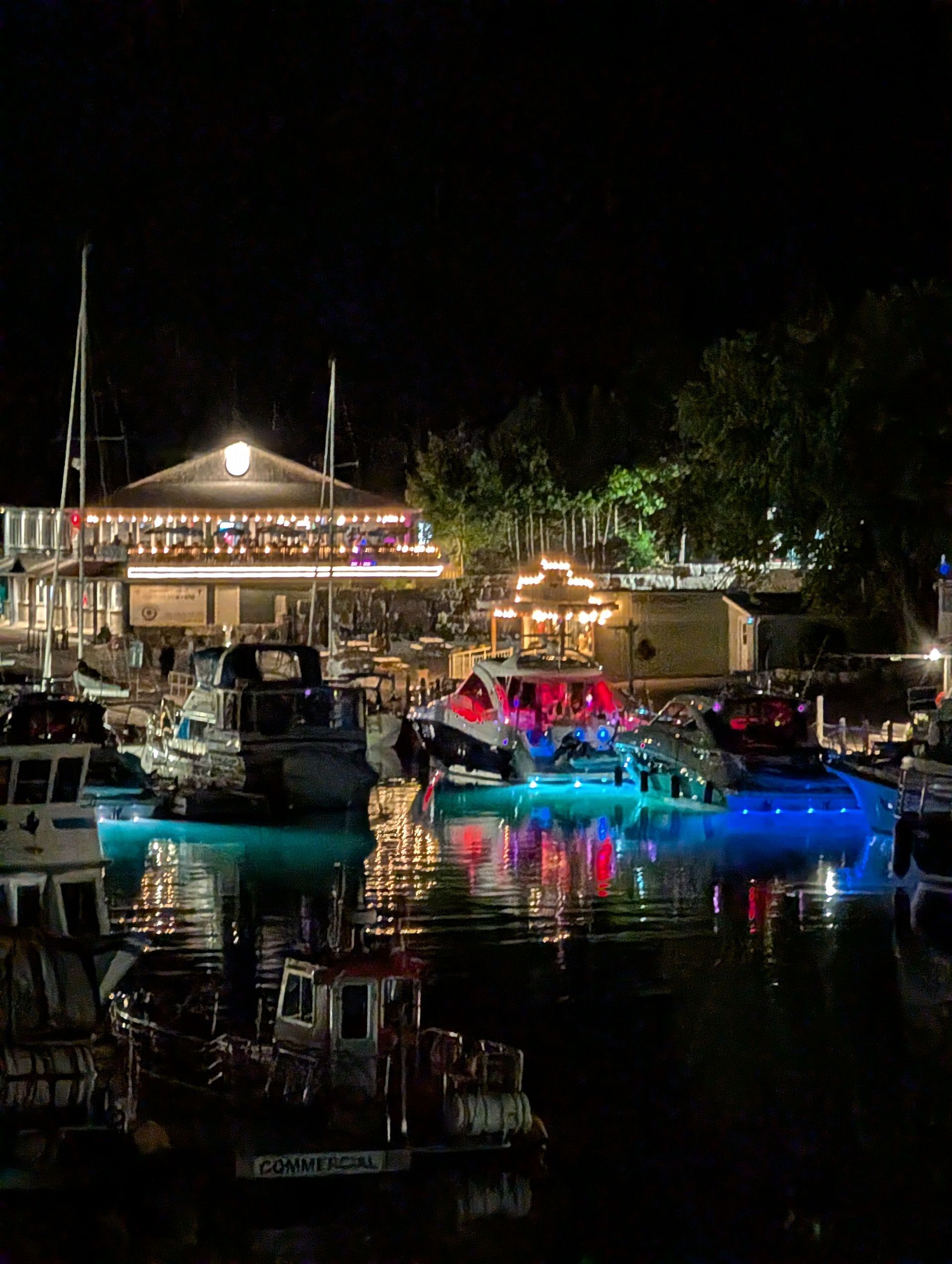 Night harbor with lit boats reflecting colorful lights on calm water near a waterfront pavilion