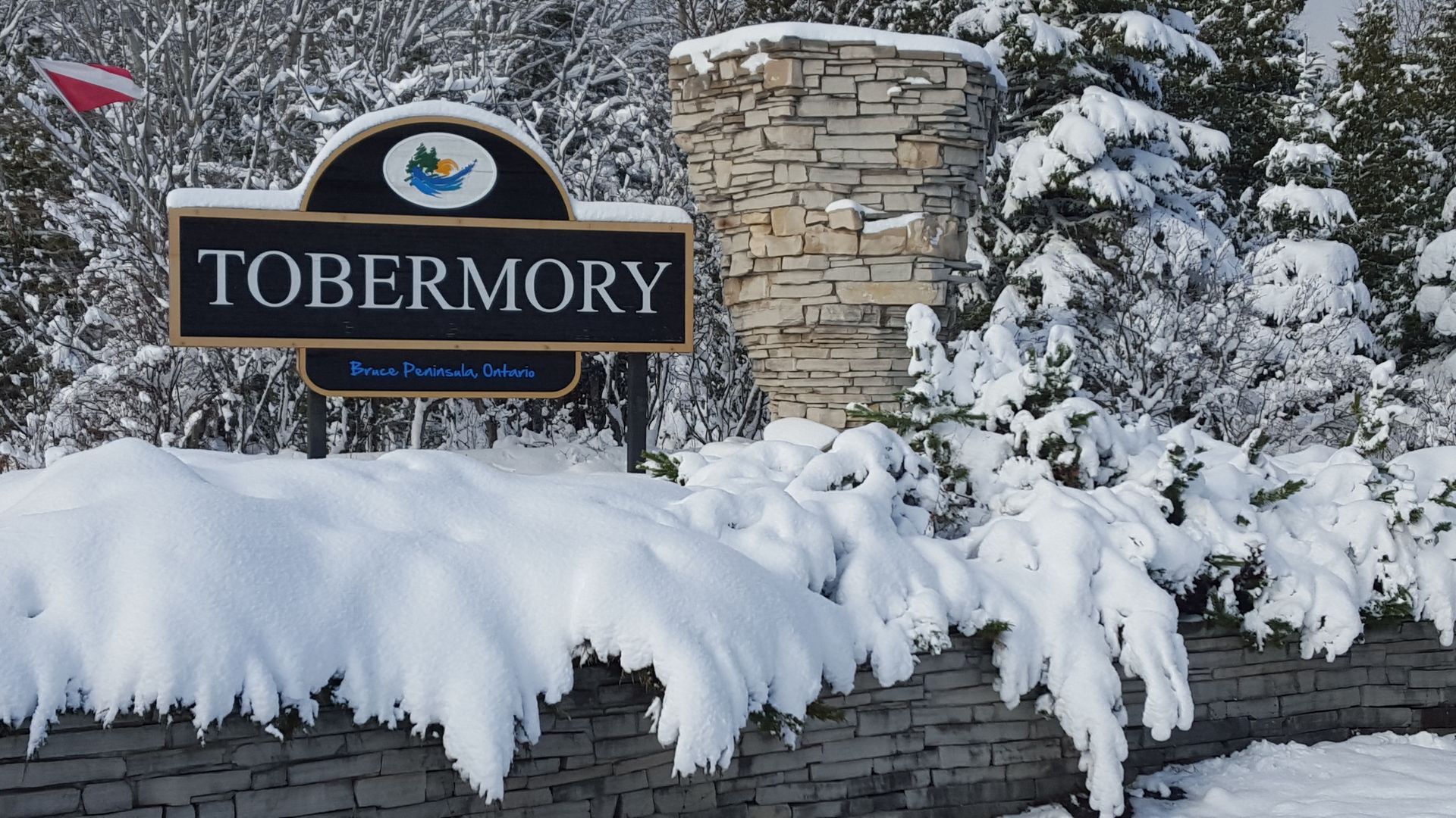 Snow-covered Tobermory town sign beside a stone wall and snowy trees