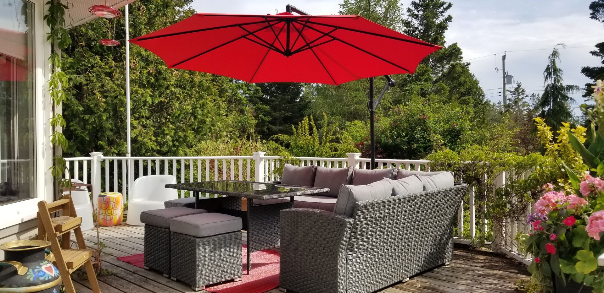 Outdoor patio with wicker seating, red umbrella, and flowers overlooking a green garden
