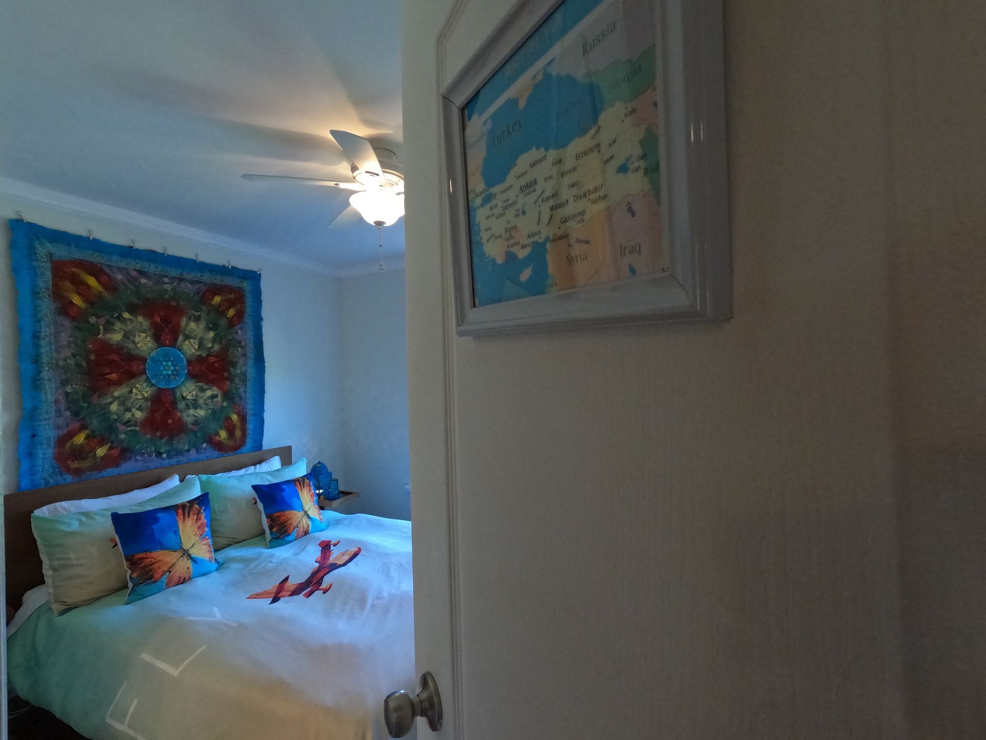 A cozy bedroom with a made bed, colorful pillows, wall art, and a ceiling fan visible through an open doorway.