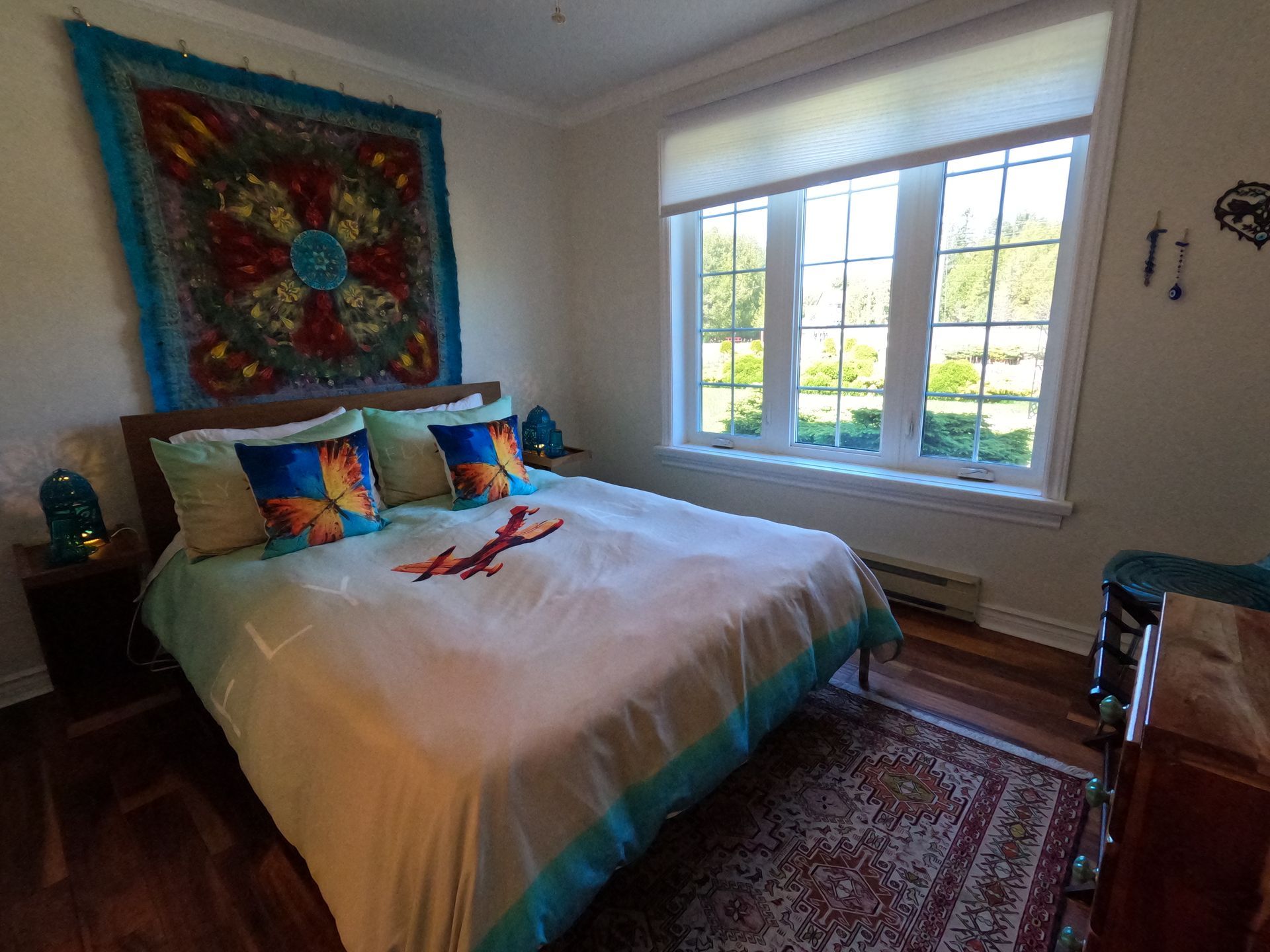 Bright bedroom with a large bed, colorful wall art, and a sunlit window over a patterned rug.