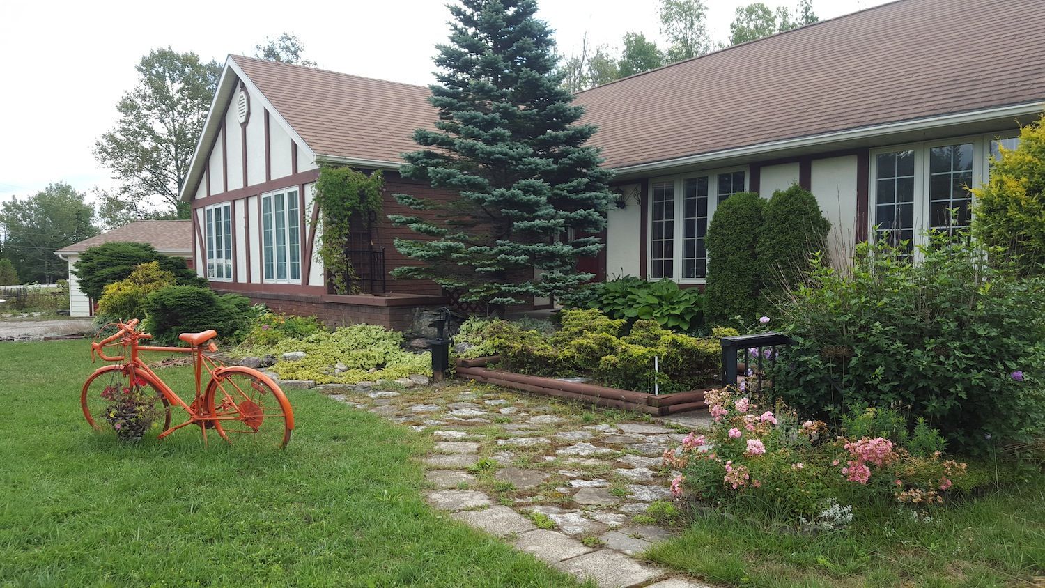 Red bicycle on grass beside a stone path leading to a brick house with a green roof and flower beds.