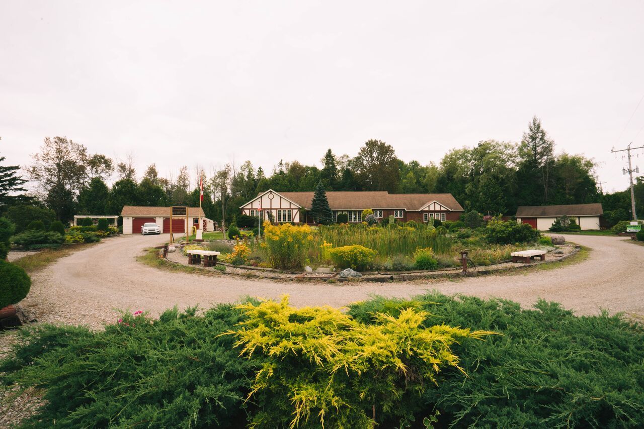 Curved gravel driveway around a flower bed and lawn in front of low buildings and trees