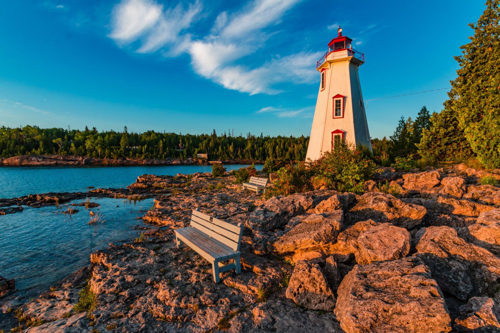 Lighthouse on rocky shoreline at sunset with a bench overlooking calm water and trees nearby