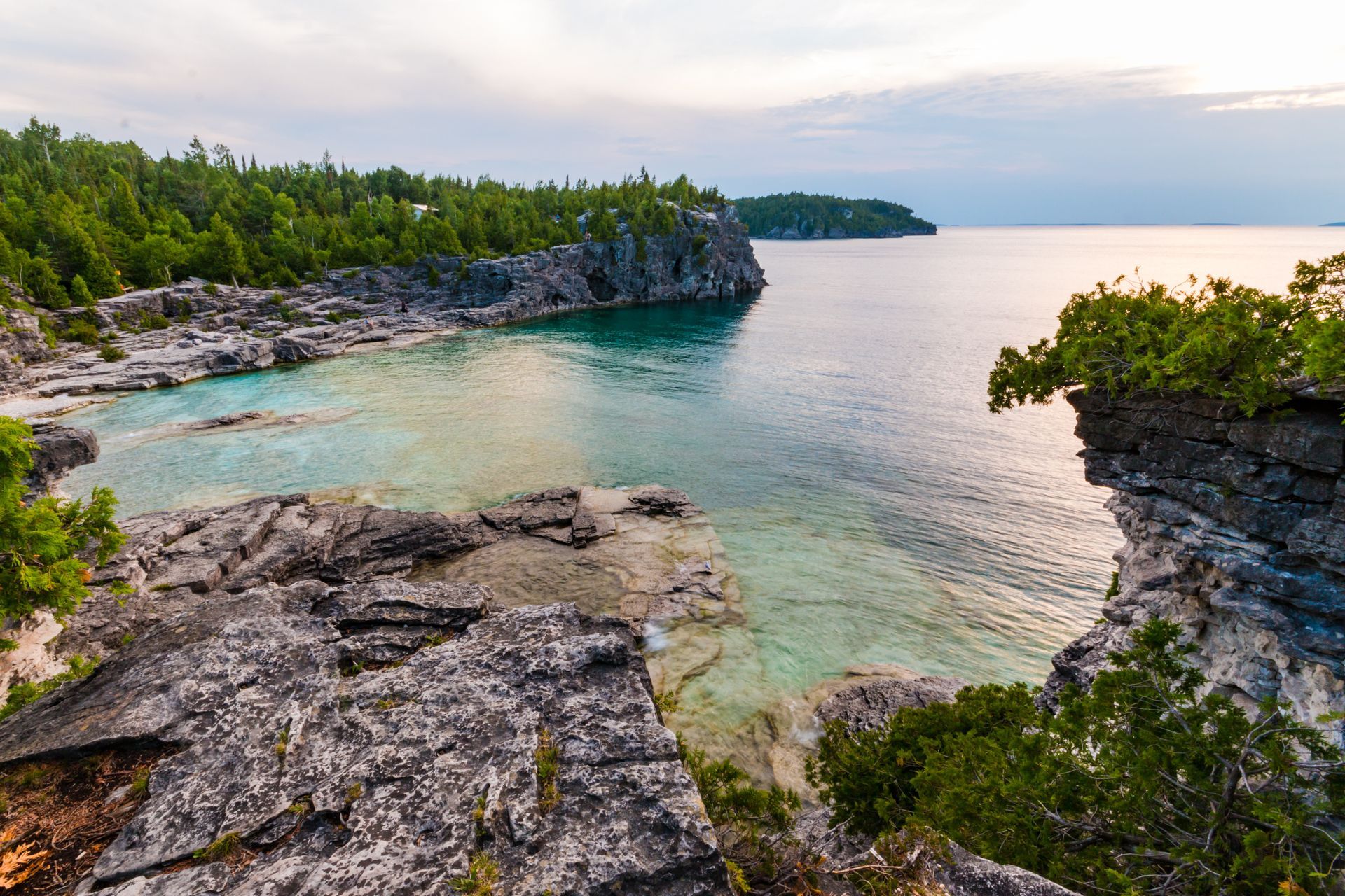 Rocky coastal cove with turquoise water, green trees, and a calm sea at sunset.
