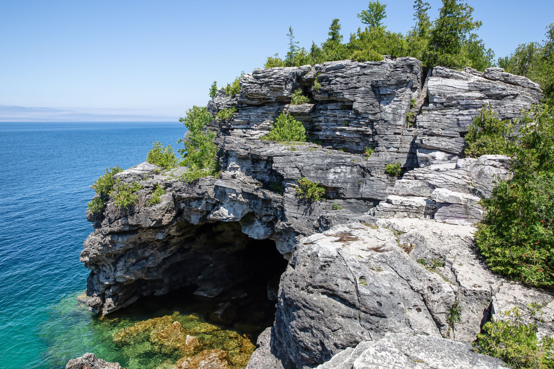 Rocky seaside cliff above clear blue water, with a small cave and green trees on top