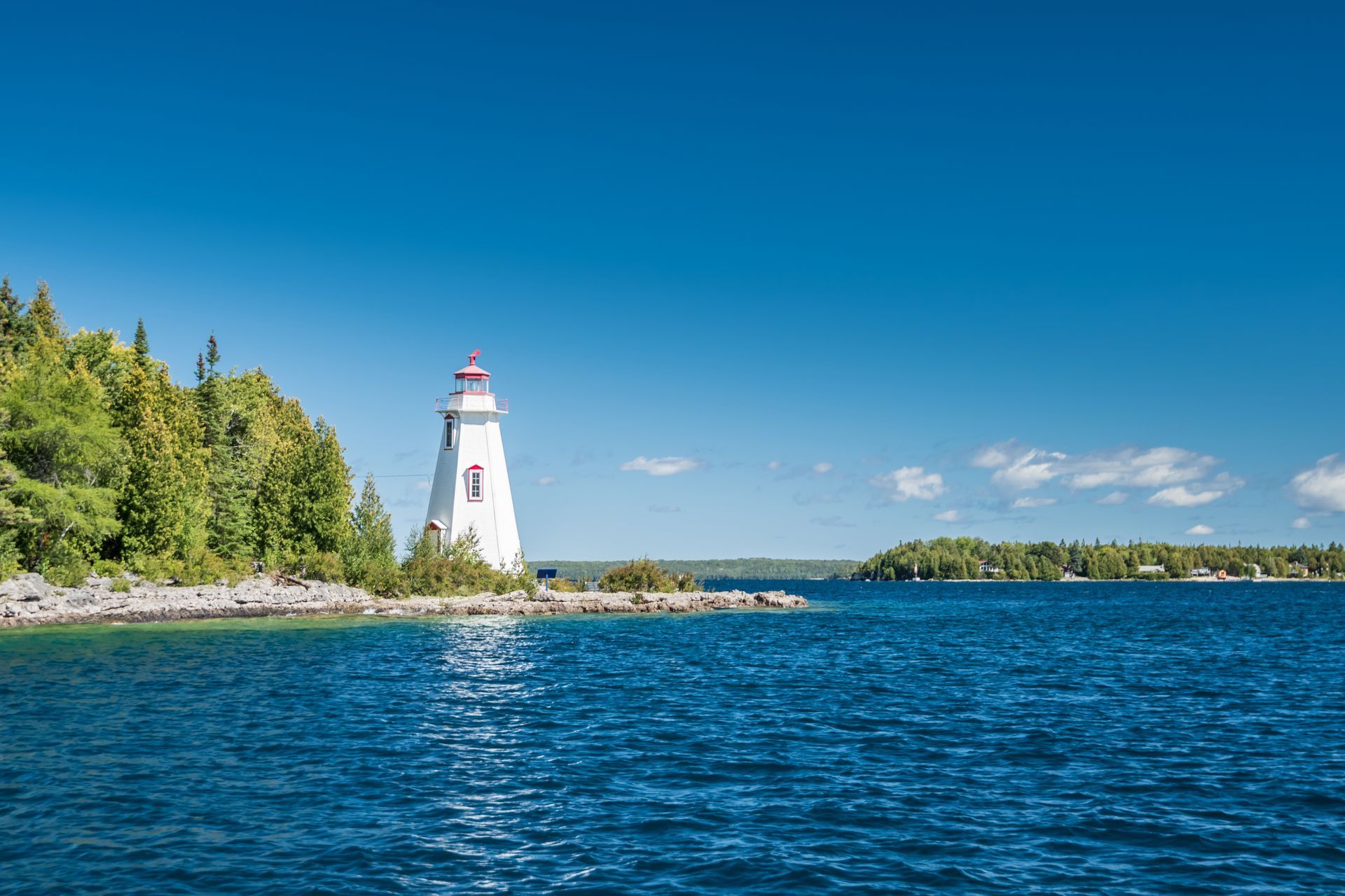 White lighthouse on a rocky shoreline beside blue water under a clear sky, with trees lining the coast